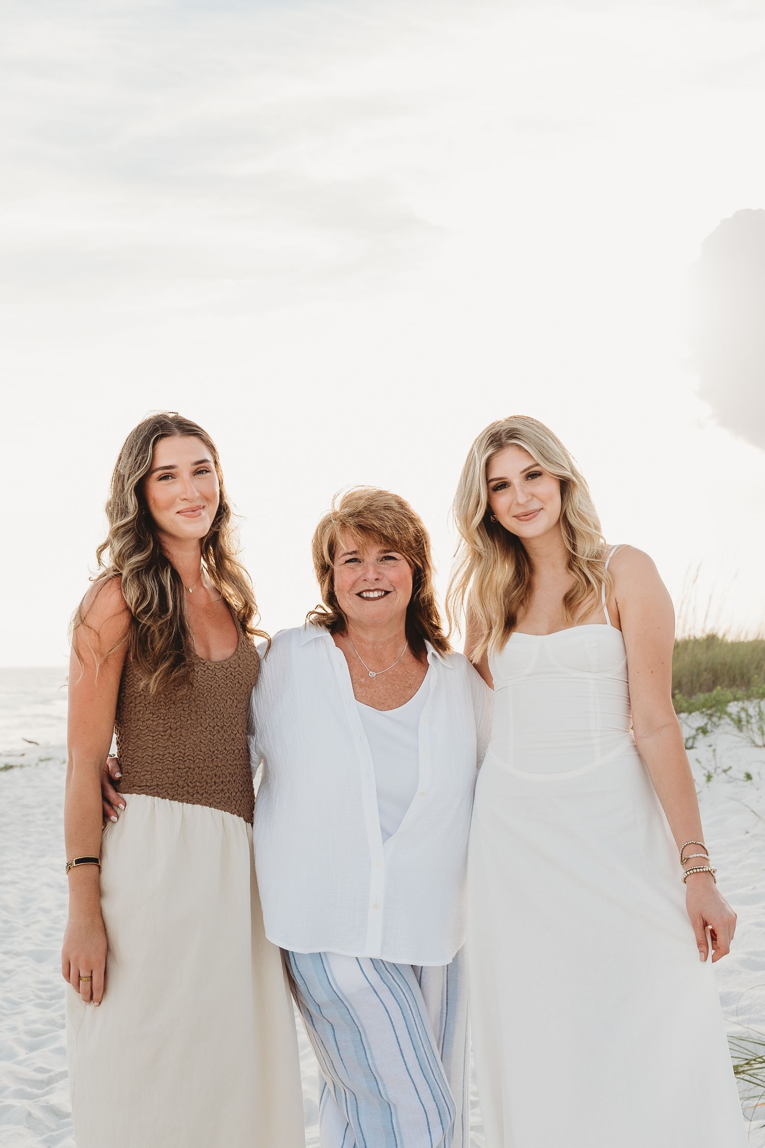 Three women pose together smiling on a beach with sand dunes and a balloon in the background.