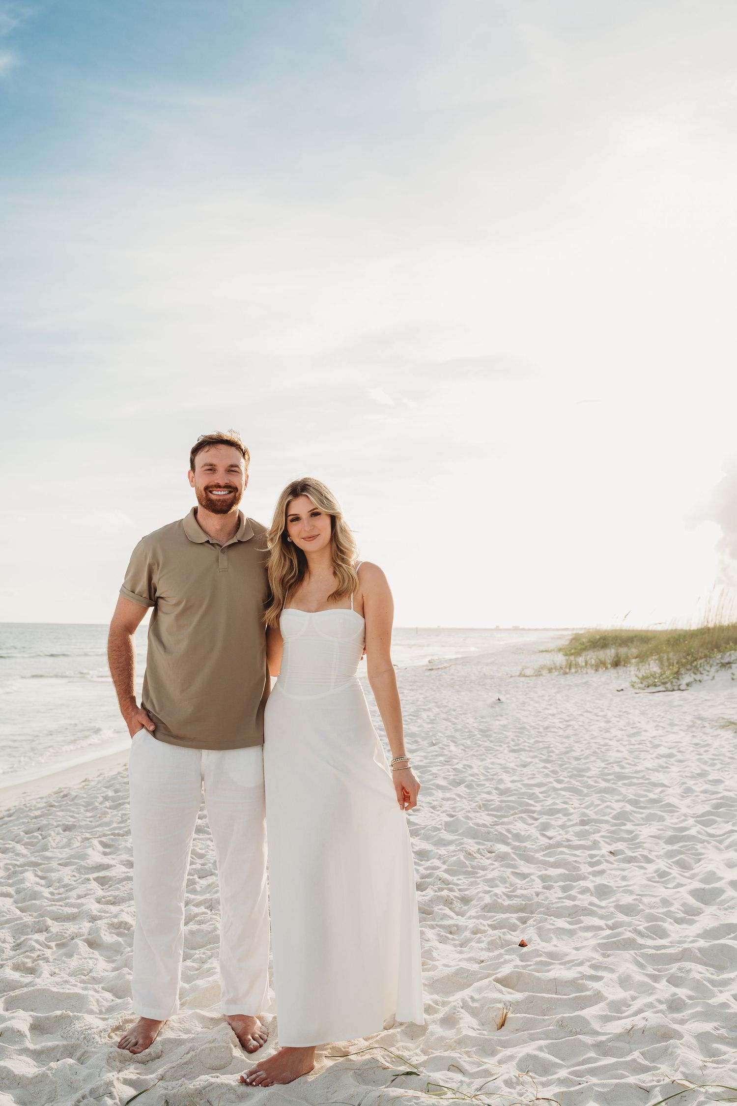 Smiling couple posing on a white sand beach, man in olive polo and woman in white dress.