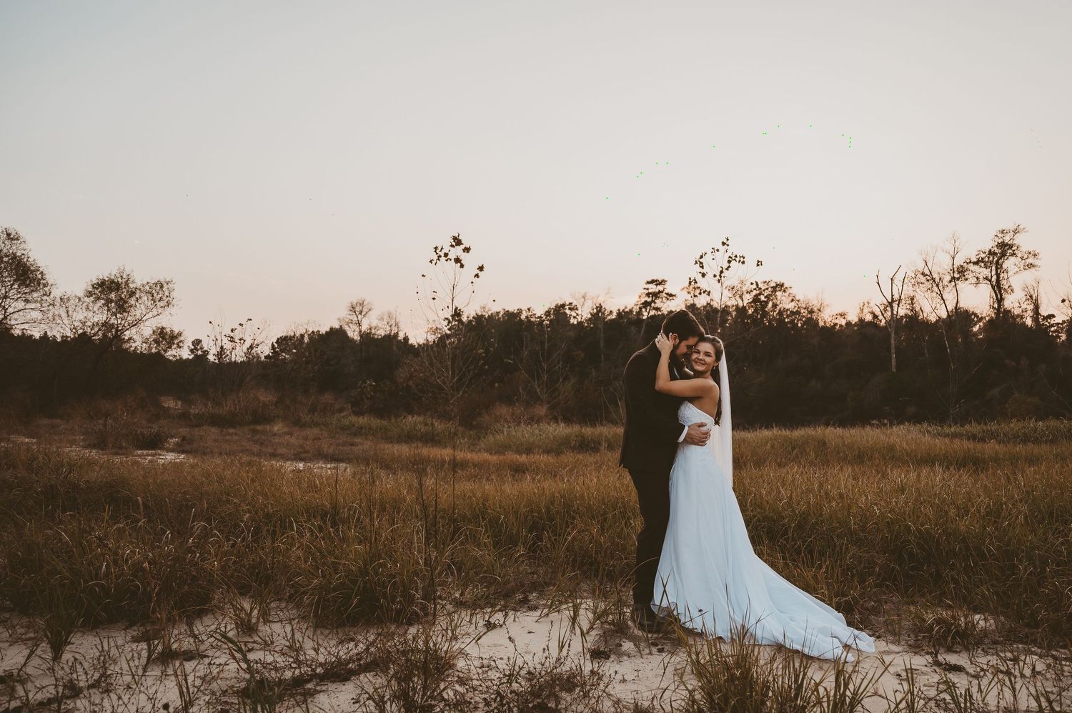 A bride and groom share a romantic embrace at sunset in a sandy field with golden grass and trees in the background.