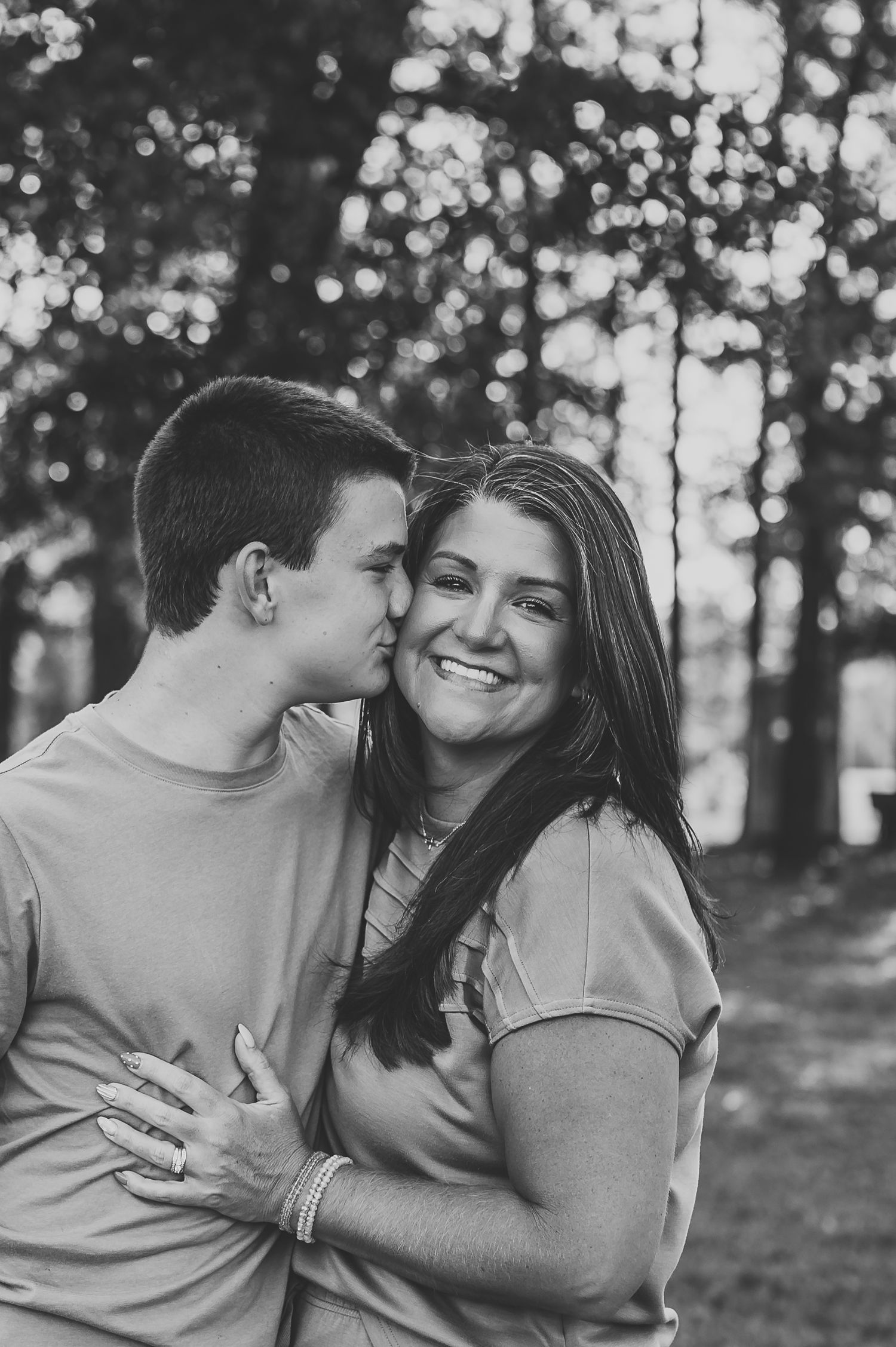 Black and white photo of a couple embracing outdoors, man kissing woman on cheek as she smiles warmly.