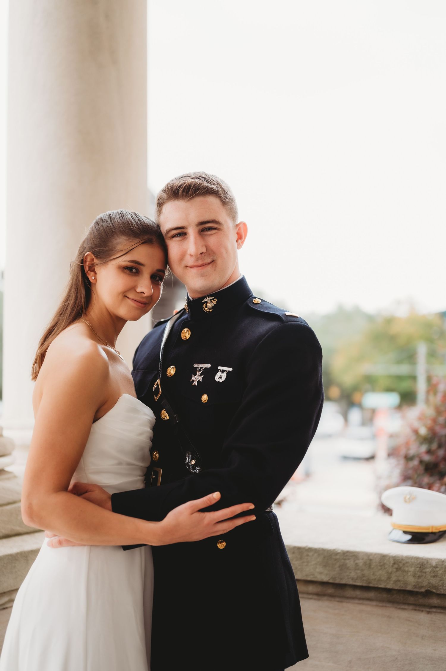 Smiling Marine groom in dress blues holds bride close between white columns.