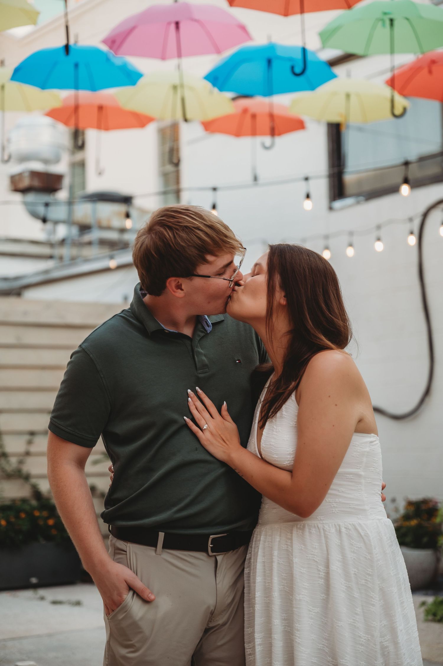 Engaged couple sharing a kiss under vibrant rainbow umbrellas with woman showing off engagement ring.