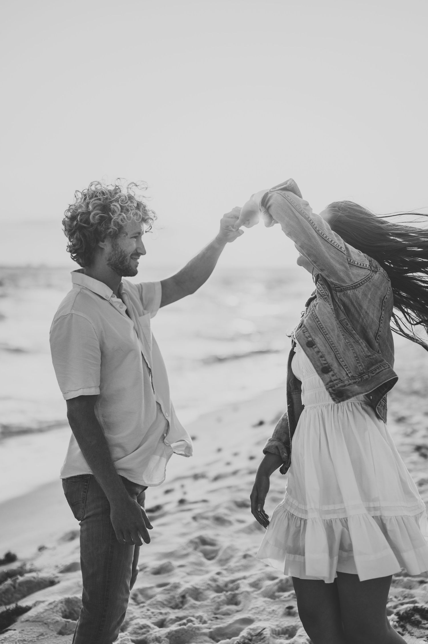 Black and white photo of curly-haired man spinning woman on sandy beach, waves in background.