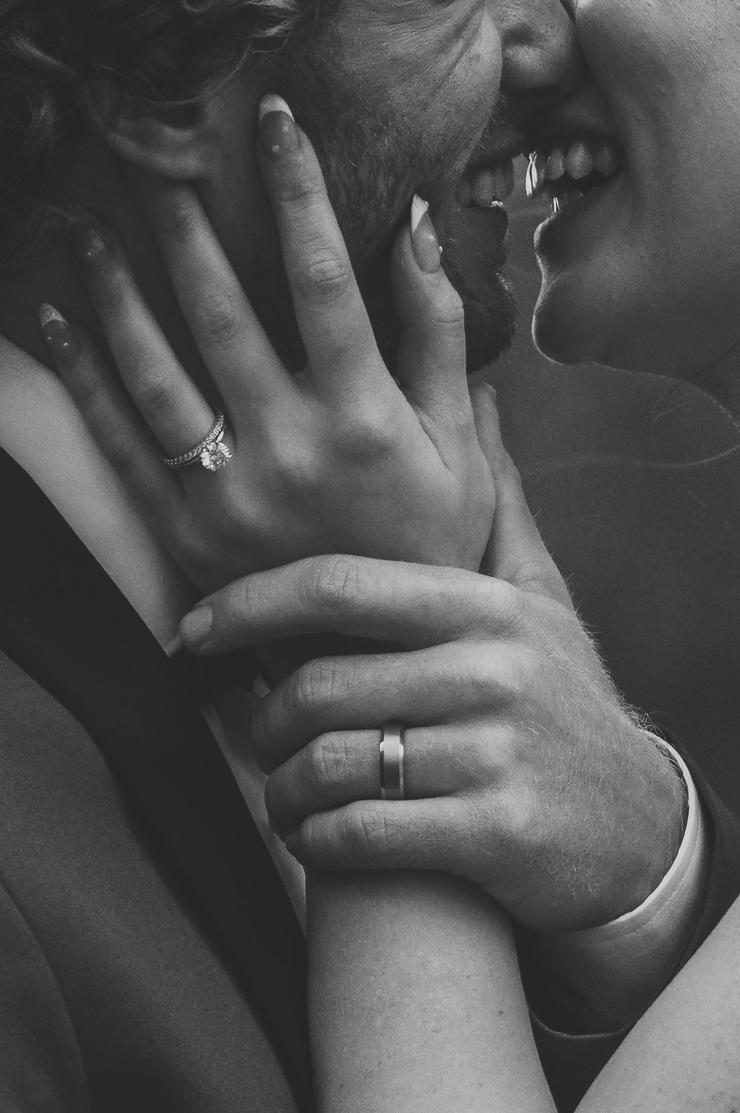 Black and white close-up of newlyweds almost kissing, showcasing engagement ring and wedding band on intertwined hands.