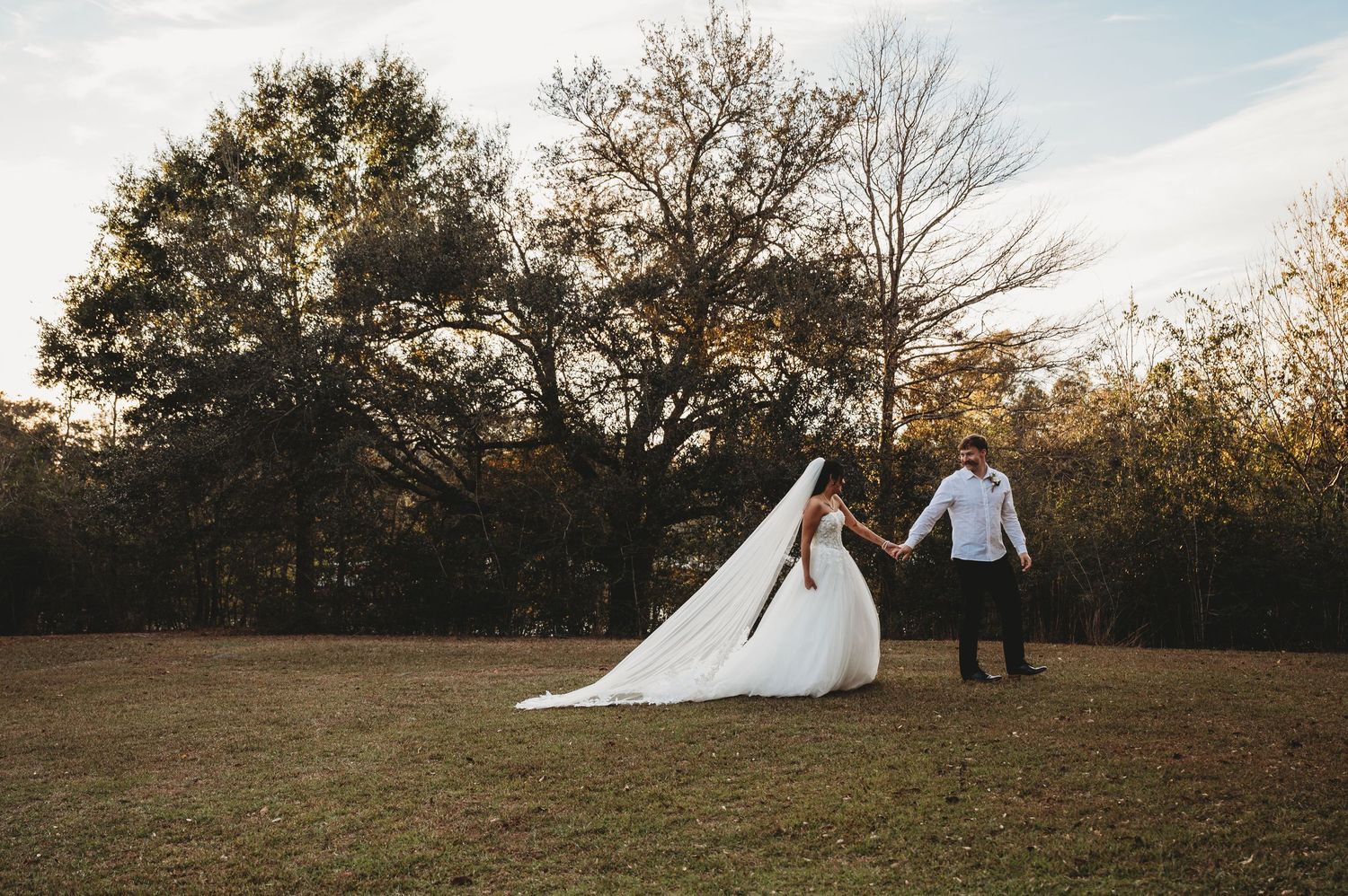 Bride and groom holding hands walking across a sunlit field with autumn trees in the background.