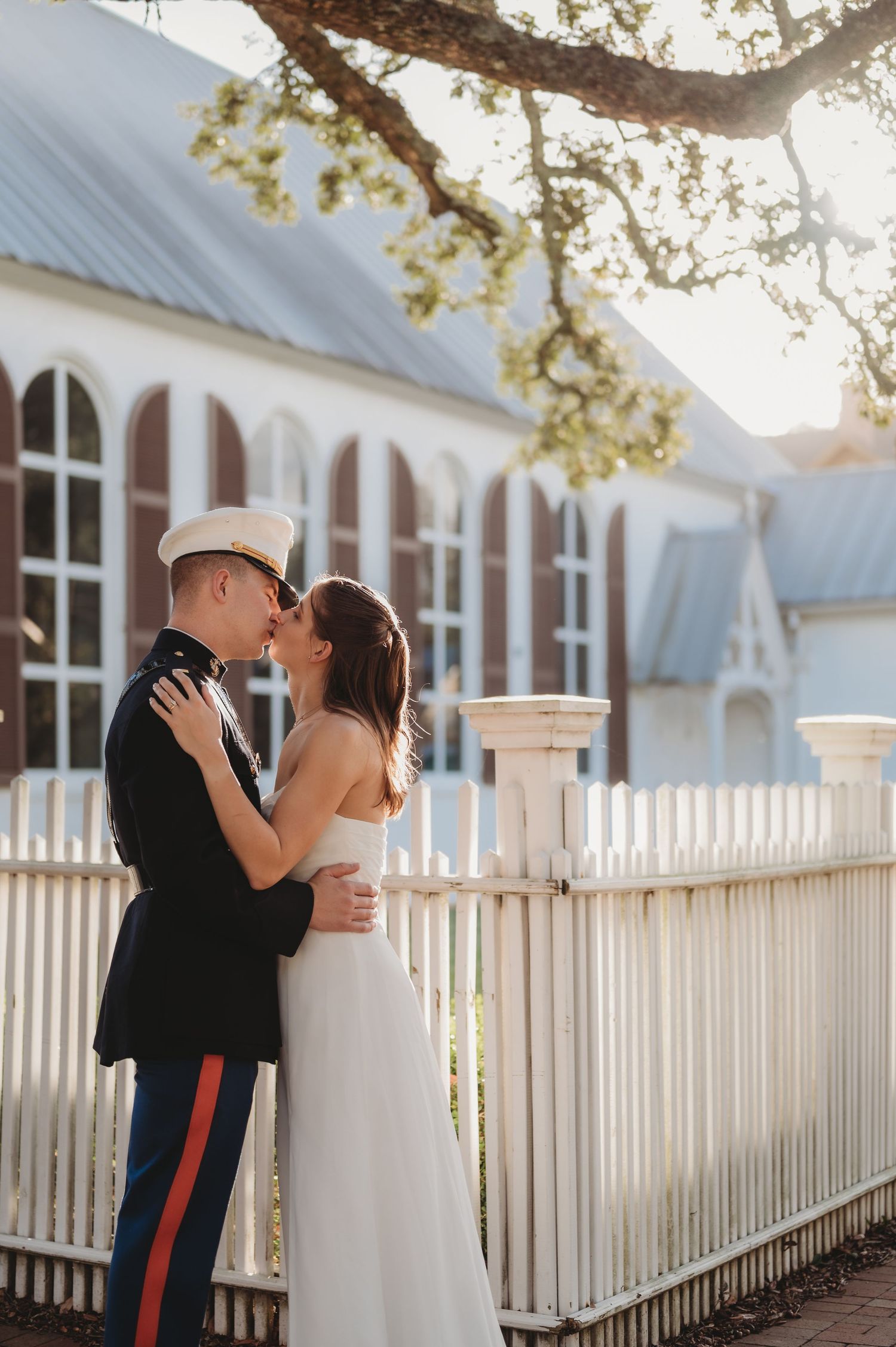 Marine groom in dress uniform kisses bride in white gown by a white picket fence outdoors.