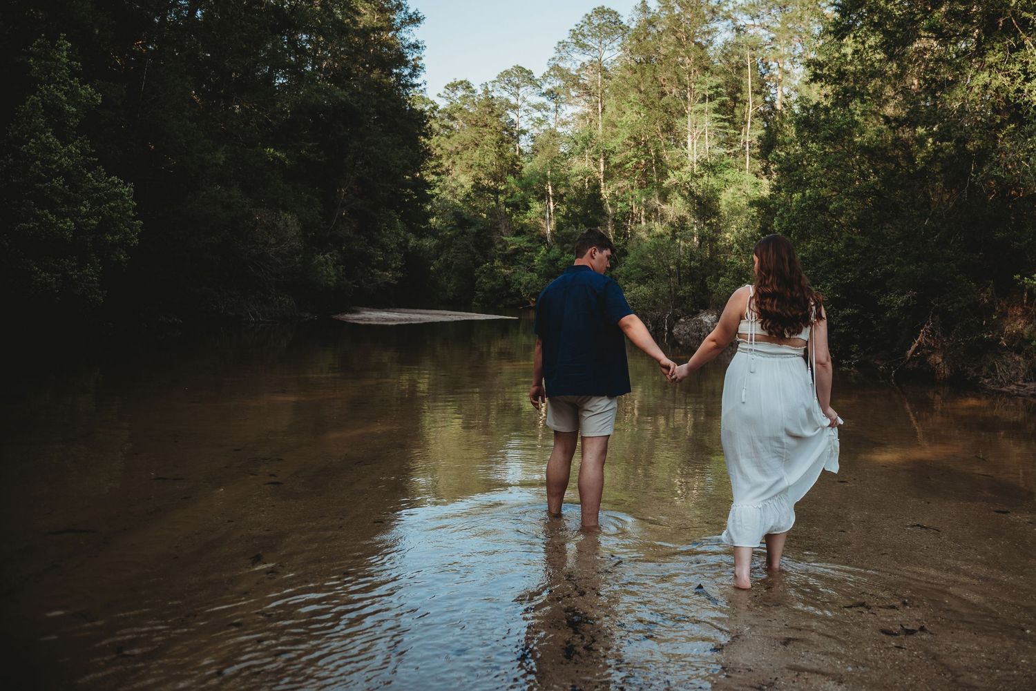 Couple holding hands walking through a shallow river surrounded by lush green forest.