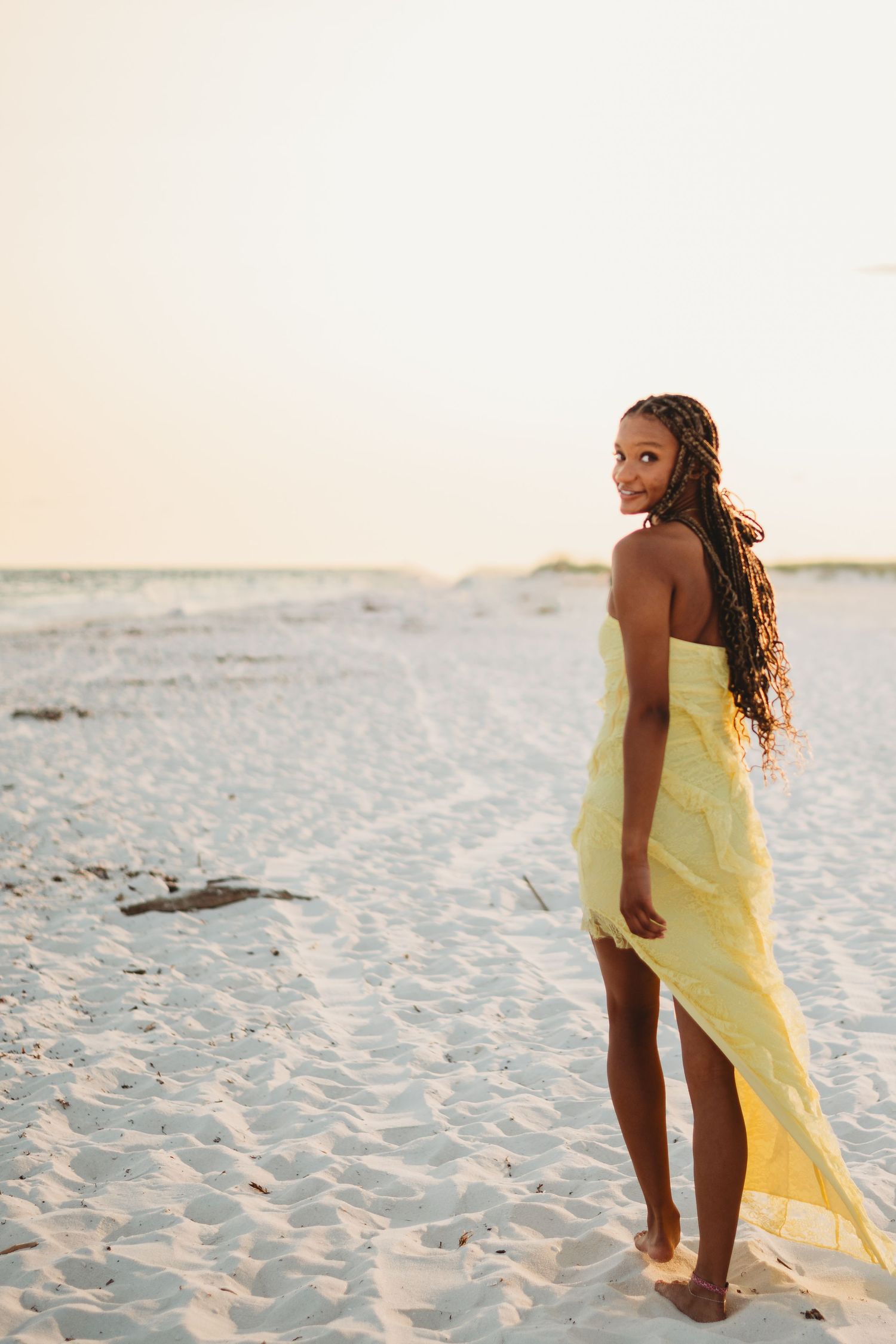 Young woman in yellow strapless dress with long braids smiling over shoulder on sandy beach at sunset.