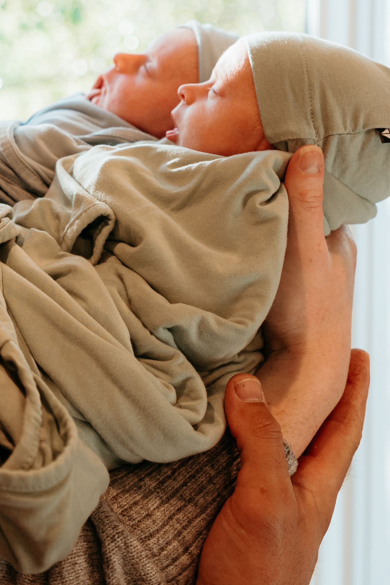 Close-up of sleeping newborn baby in sage green outfit and white hat cradled in parent's hands.