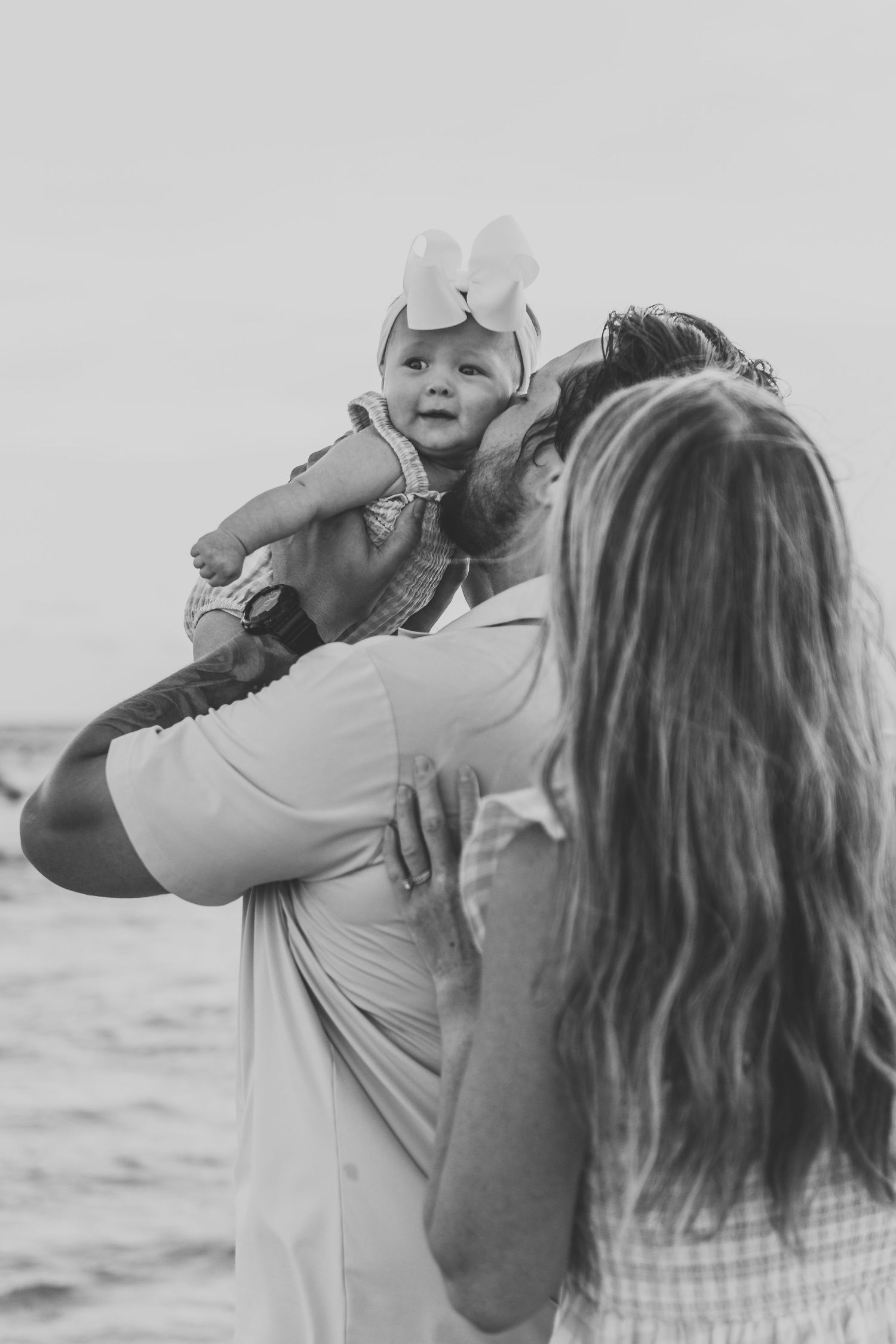 Black and white photo of parents lifting their baby girl with a bow headband at the beach.