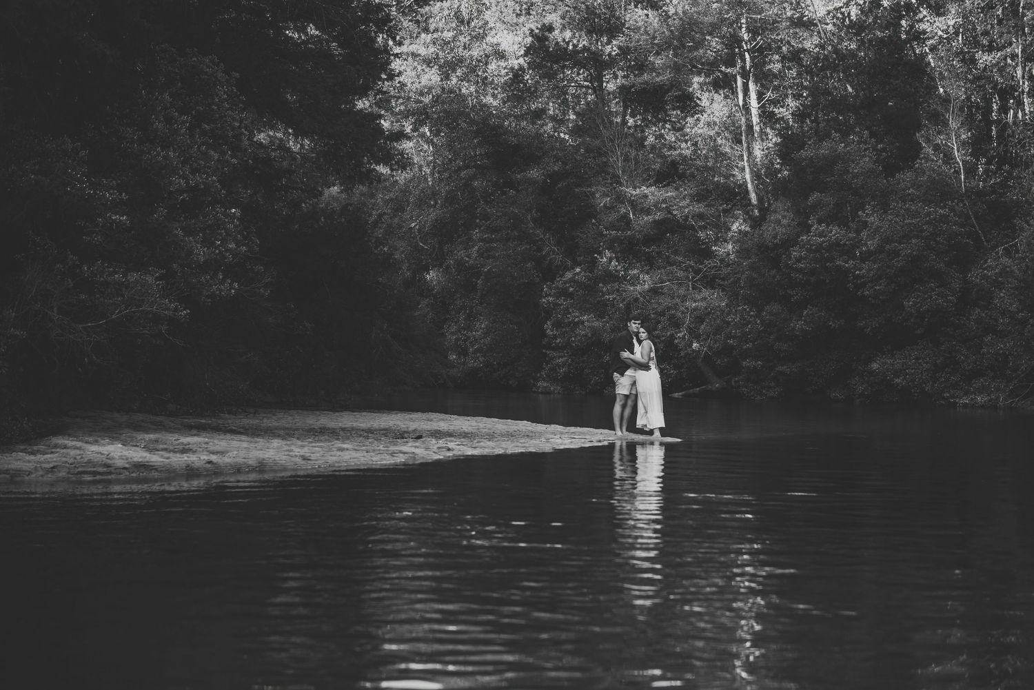 Black and white photo of couple embracing on a sandbar in a peaceful forest river.