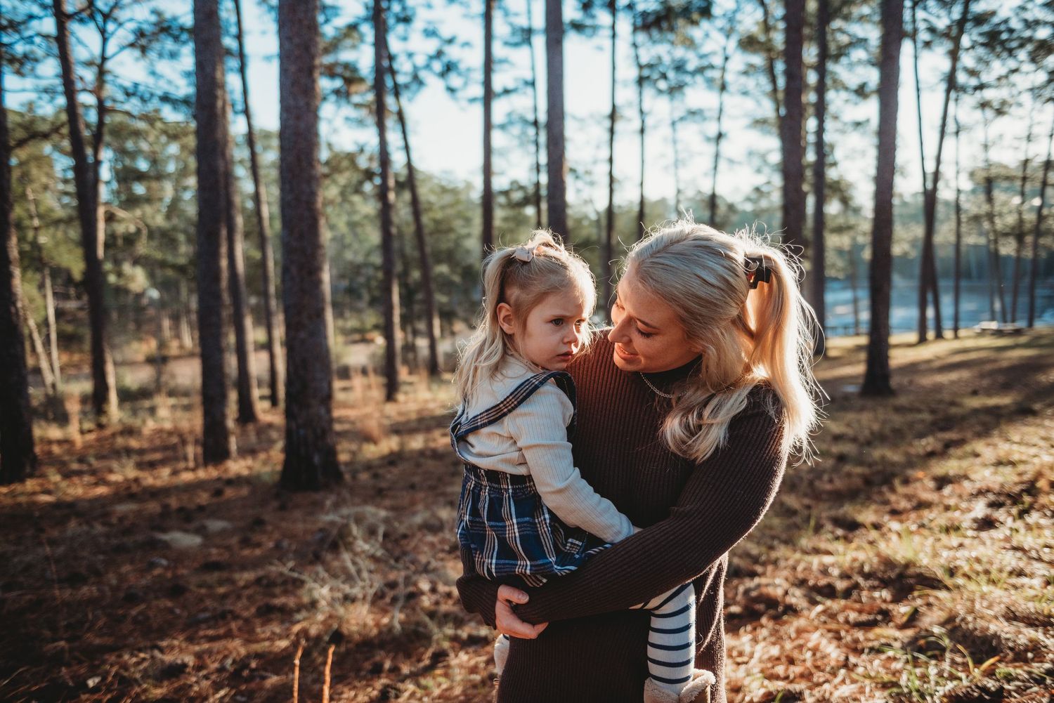 Mother holds young daughter in sunlit pine forest, sharing a tender moment in warm natural light.