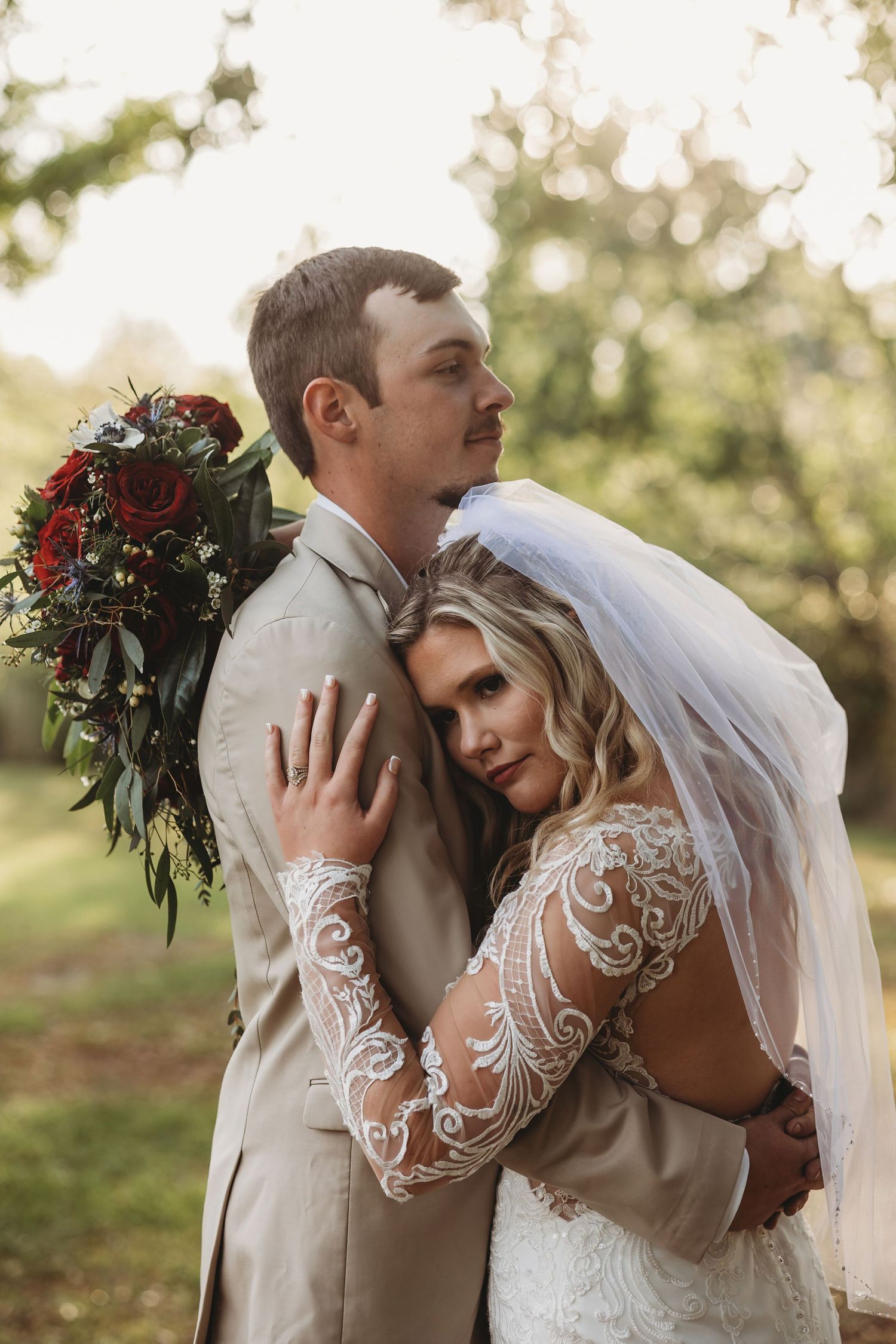 Bride in lace gown embraces groom in tan suit outdoors, holding red rose bouquet during golden hour wedding portrait.