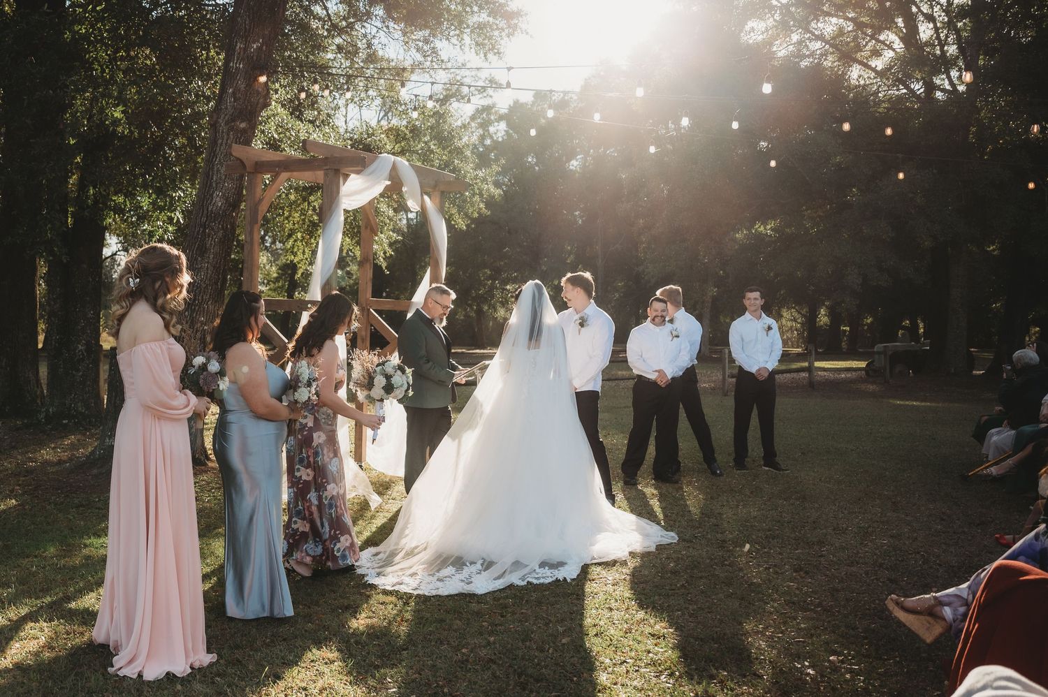 Outdoor wedding ceremony under arch with bridal party in sunlit forest setting.