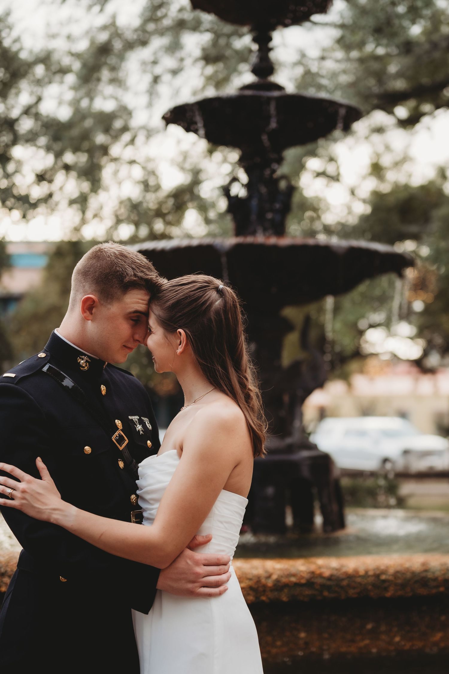 Newlywed couple embraces intimately in front of ornate black fountain, groom in Marine dress uniform.