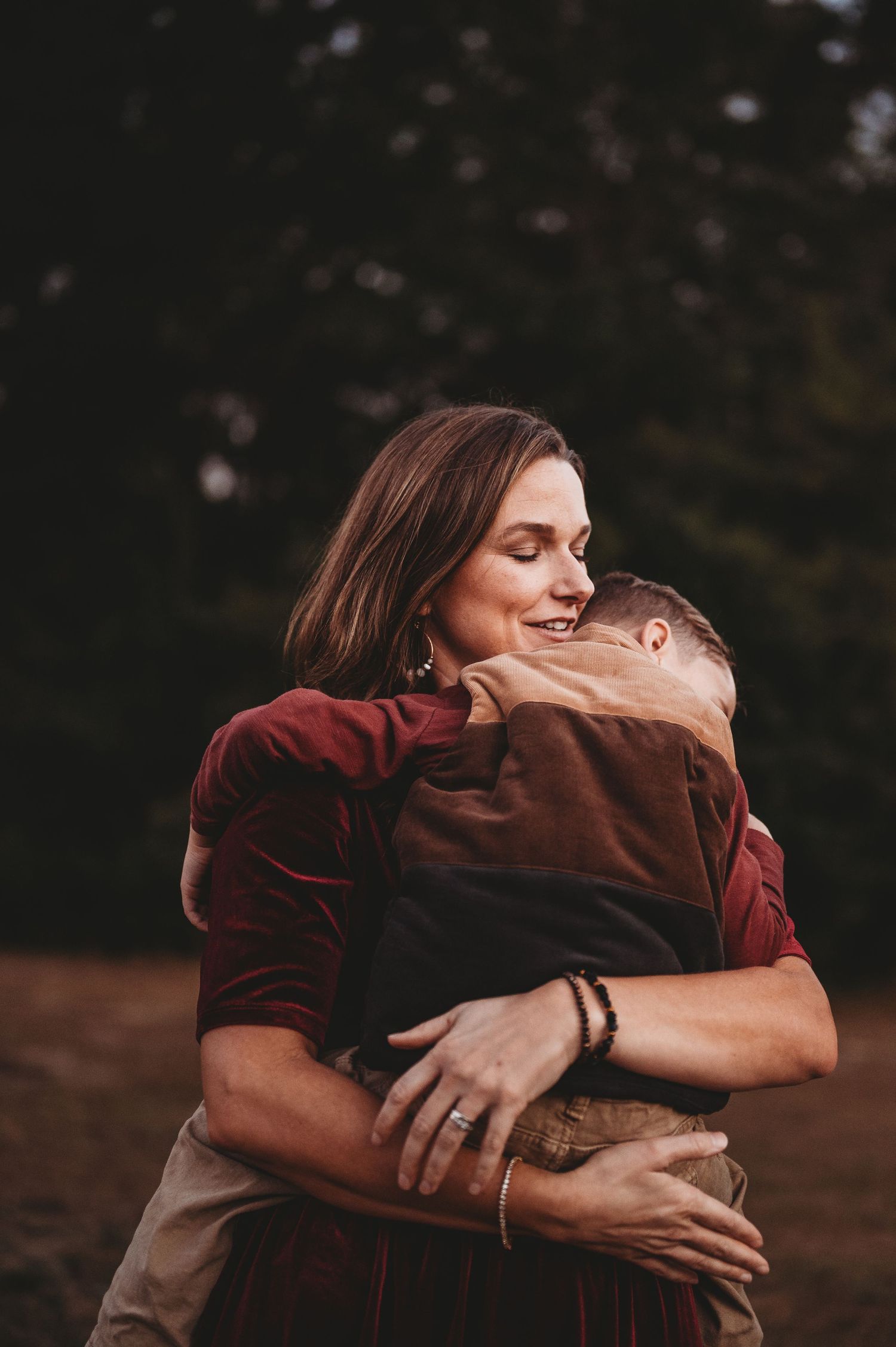 A smiling woman with brown hair joyfully hugs a child outdoors, wearing a red shirt and bracelet.