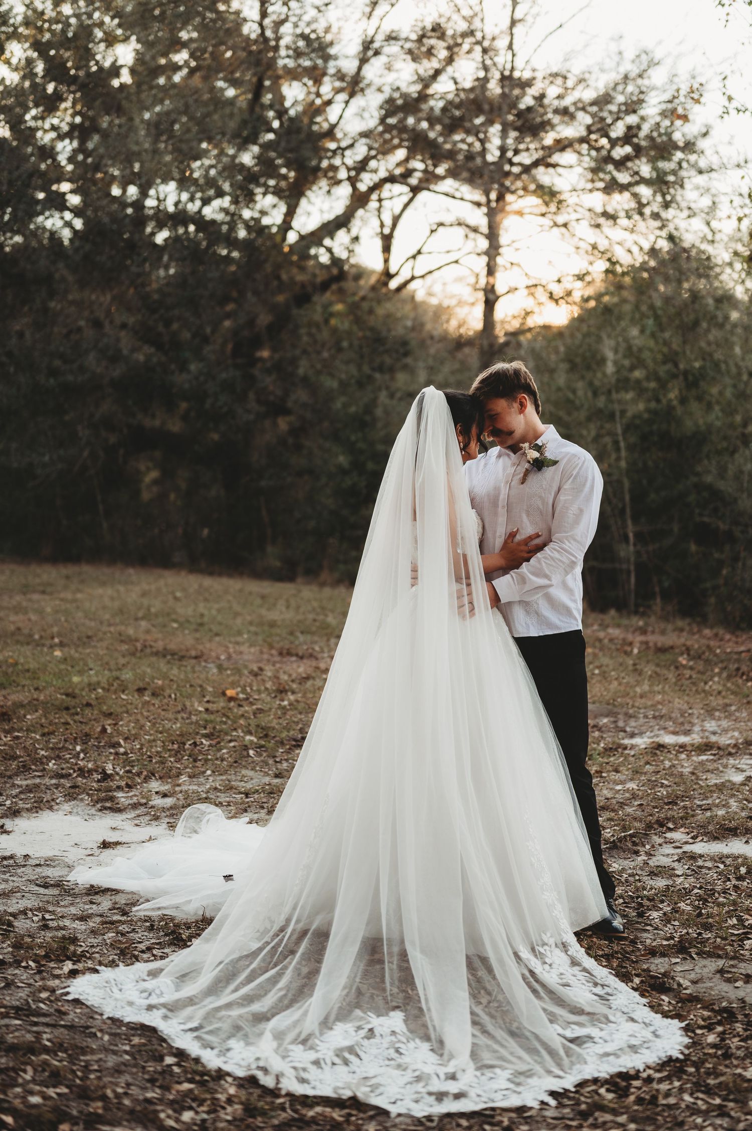 Bride and groom sharing an intimate moment outdoors at sunset, with a flowing cathedral veil trailing on the ground.