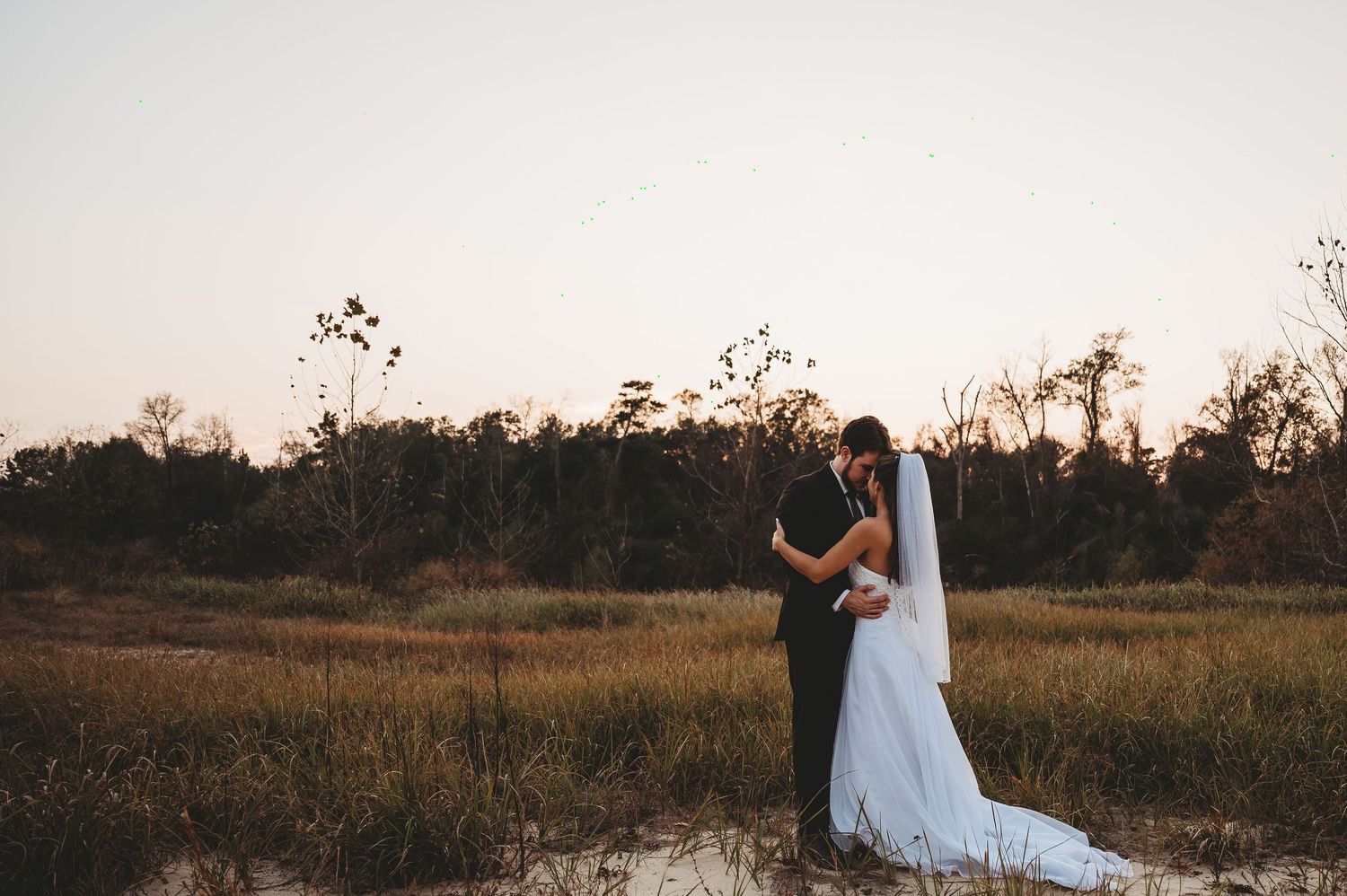 Bride and groom embracing at sunset in a golden field with trees in the background on their wedding day.