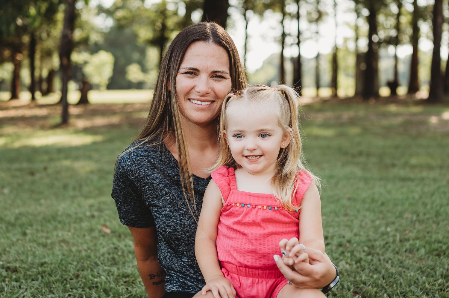 A smiling woman holds a young blonde girl in a pink outfit outdoors in a green park setting.
