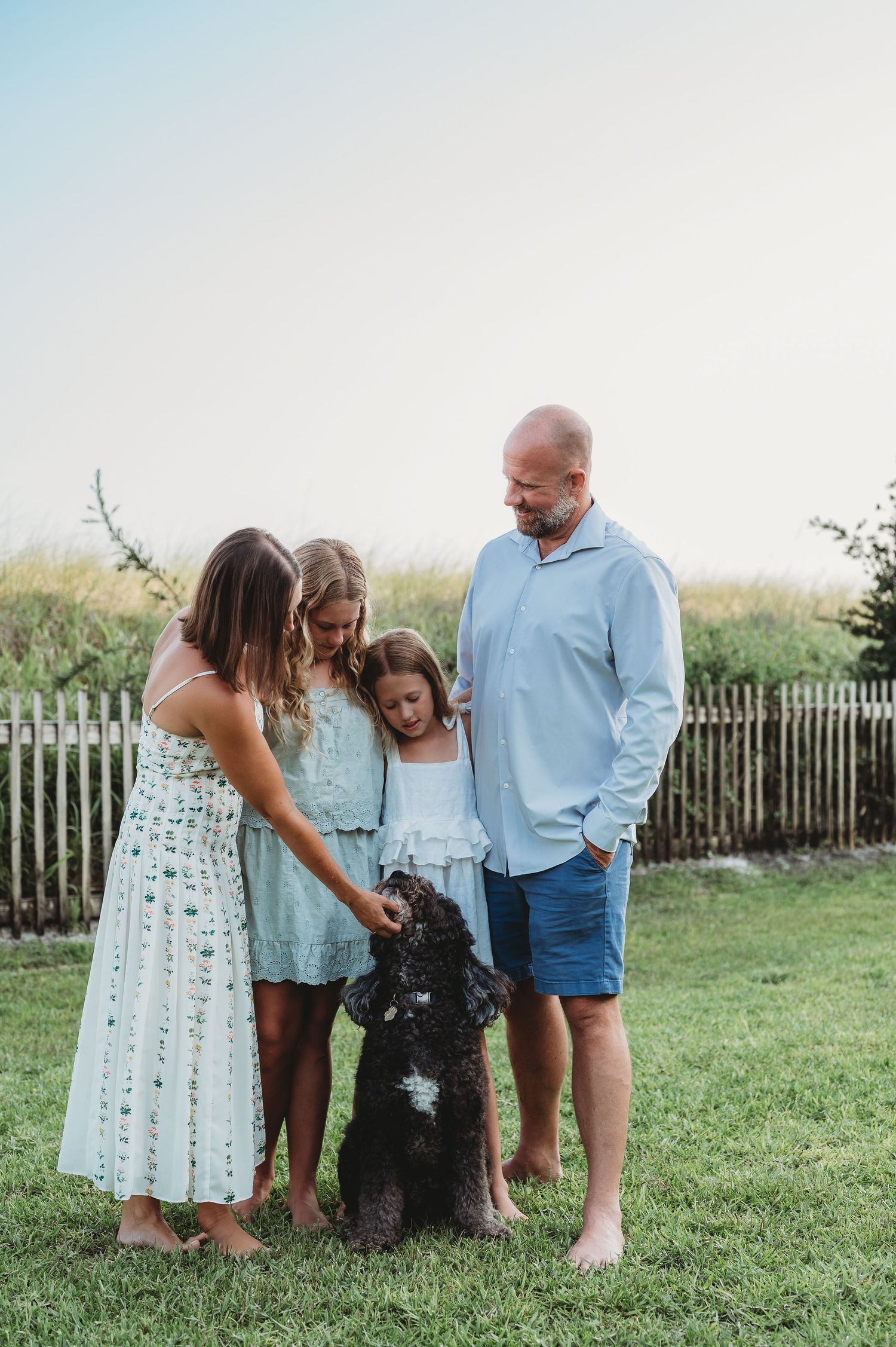 Family of four with two girls petting a black dog on a green lawn near a white fence outdoors.
