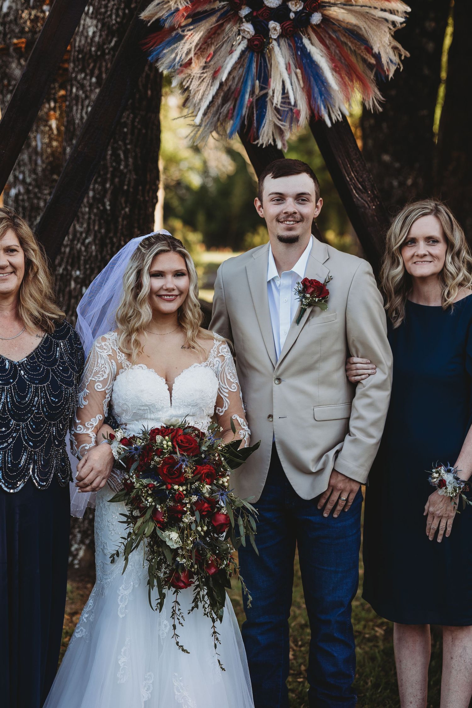 Bride in lace gown with red rose bouquet and groom in tan suit posing with family outdoors.
