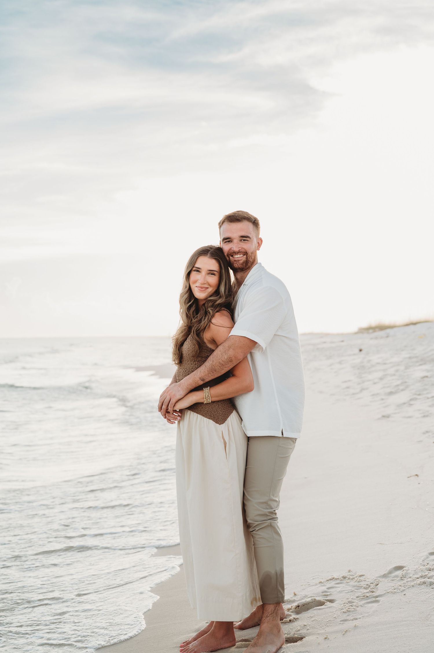 A couple embraces on a sandy beach at sunset, man in white shirt hugging woman in neutral dress.