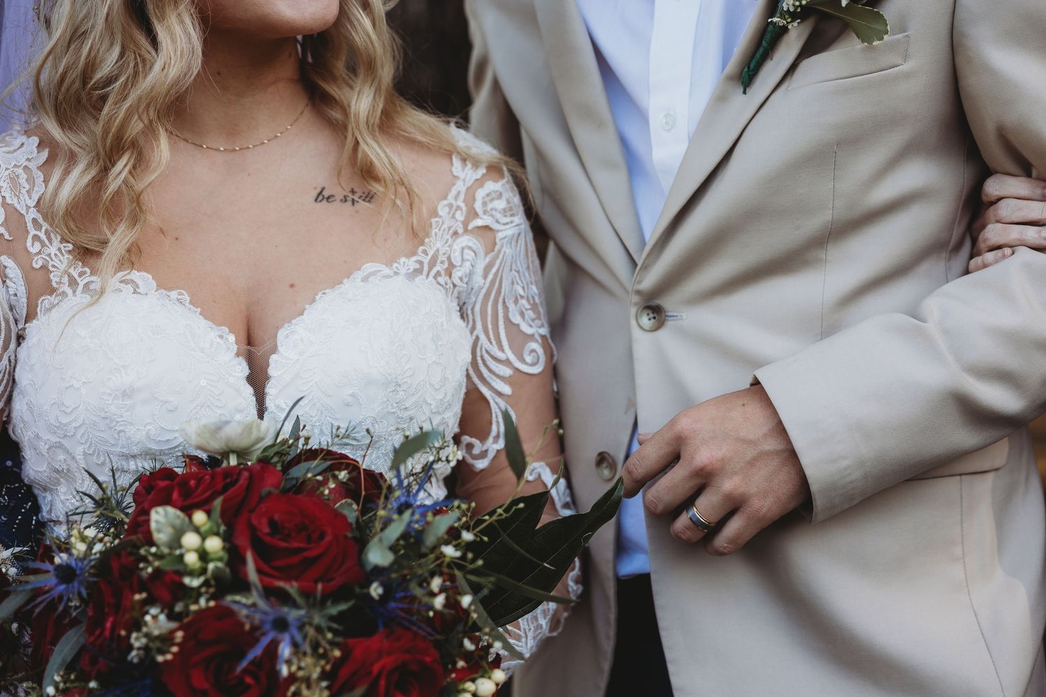 Close-up of bride in lace gown holding red rose bouquet standing beside groom in tan suit.