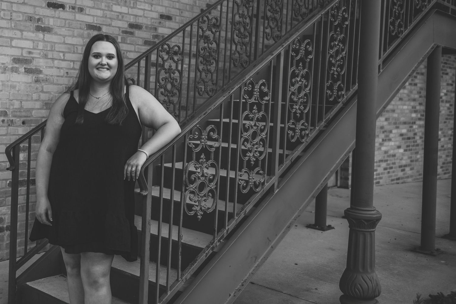 A woman in a black dress poses on an ornate iron staircase in black and white.