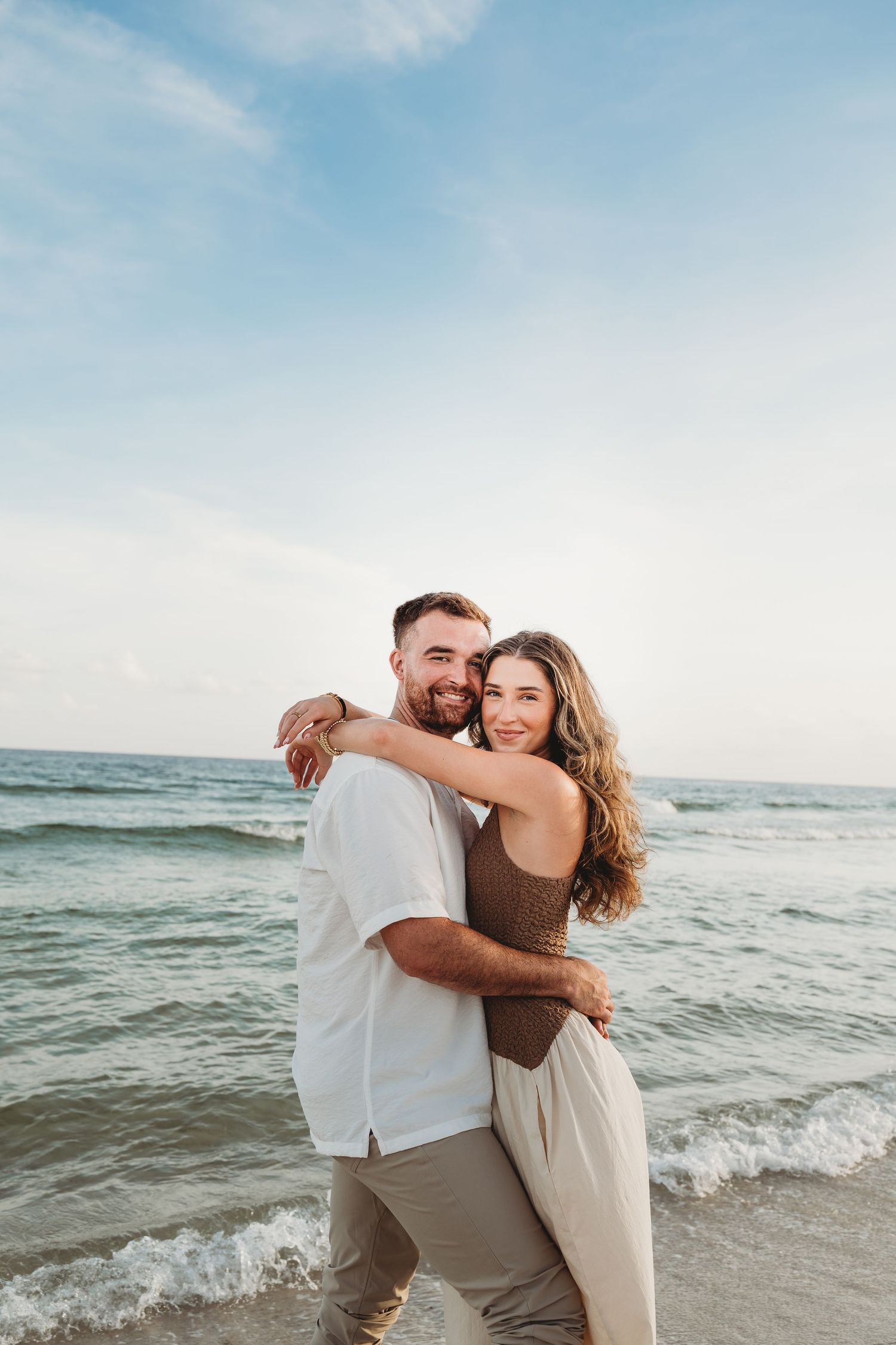 A couple embraces and smiles on a beach with ocean waves and blue sky in the background.
