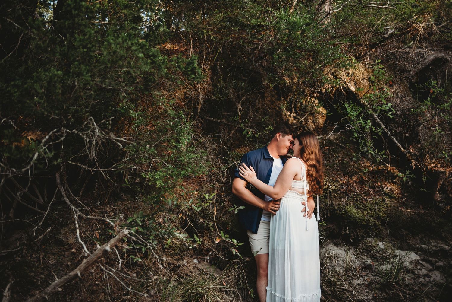 Couple embracing tenderly among dense forest greenery and rocky terrain in soft natural light.
