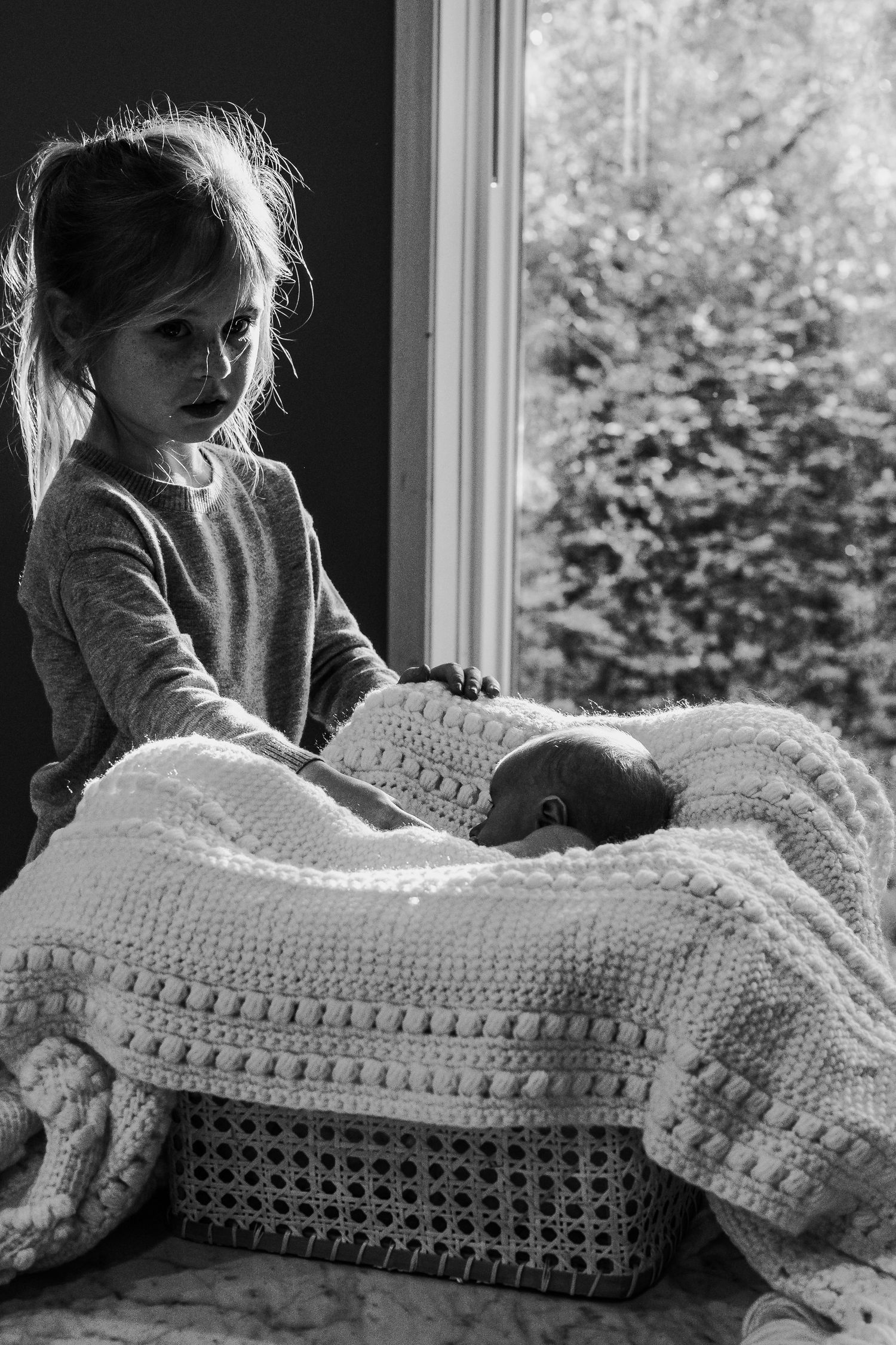 Black and white photo of a young girl lovingly watching over twin newborns in a wicker bassinet by a window.