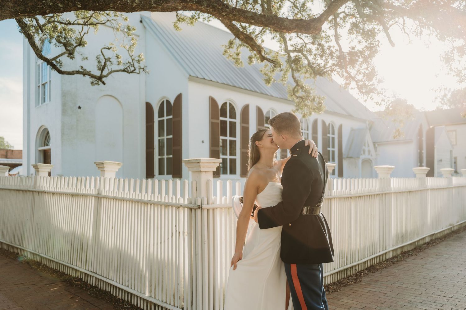 Military groom and bride share a romantic kiss outside a white church with picket fence at golden hour.