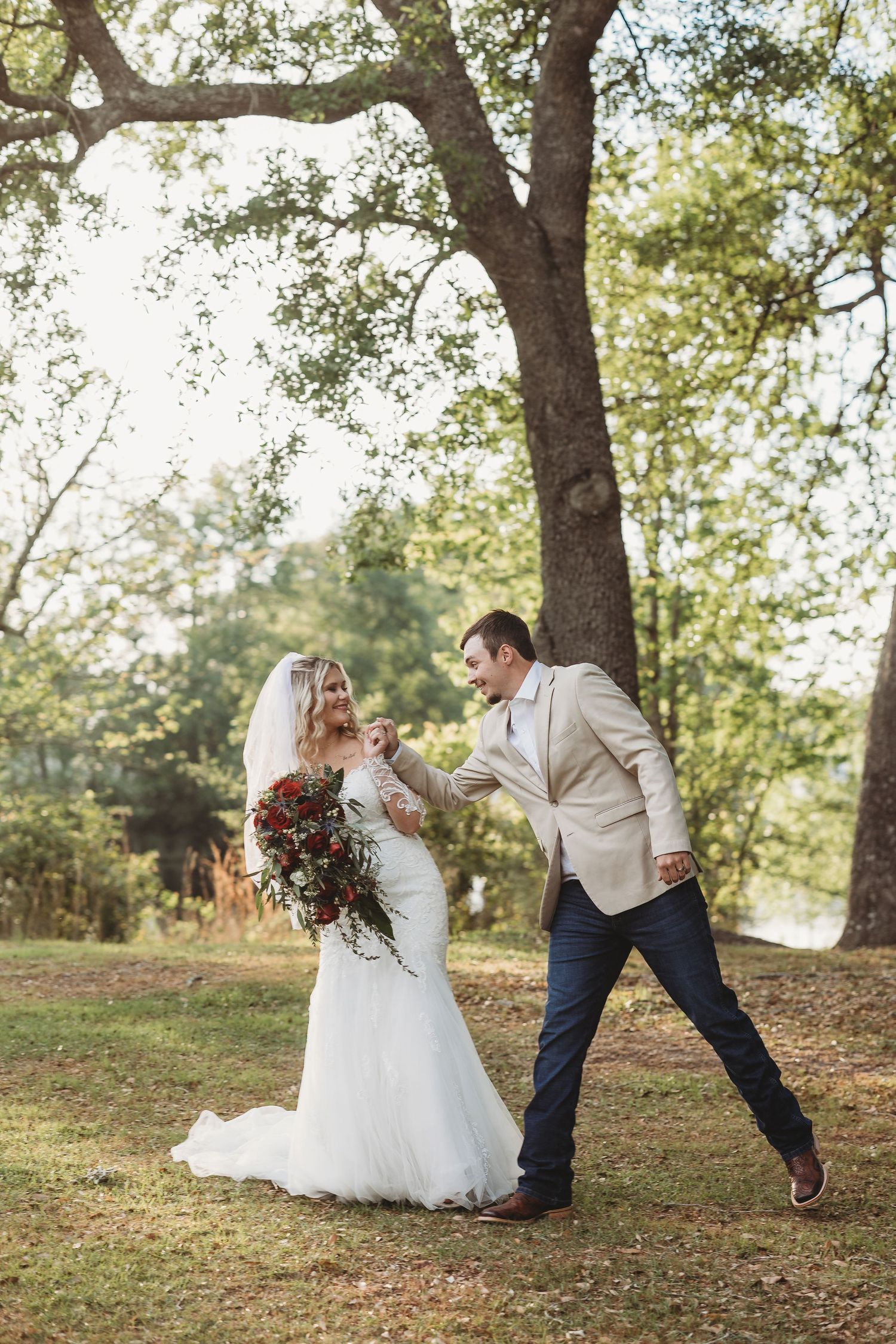 Bride and groom dancing joyfully outdoors under large oak trees, bride holding red bouquet in white gown.