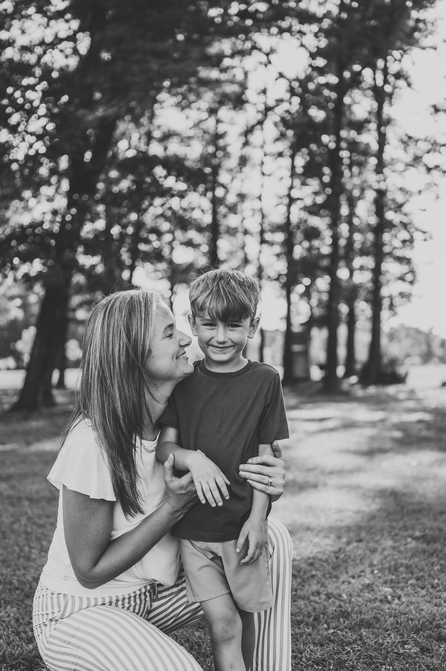 Black and white photo of a smiling mother hugging her young son outdoors in a park surrounded by tall trees.