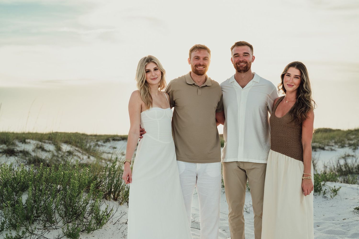Two couples smiling together on a white sand beach at sunset.