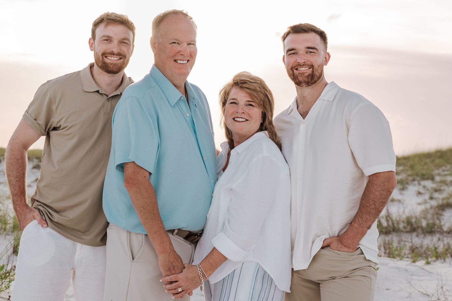 Family of four smiling together on a beach at sunset near sand dunes in casual attire.