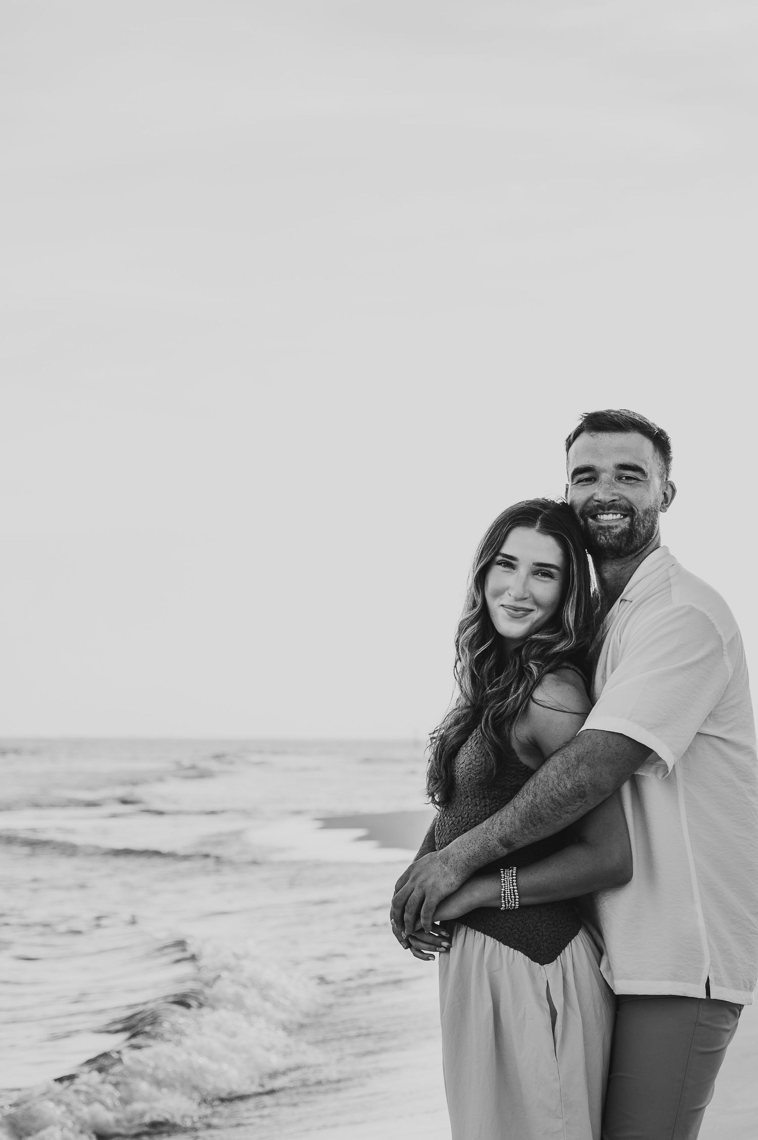 Black and white photo of a man embracing a woman from behind on a beach by the ocean.