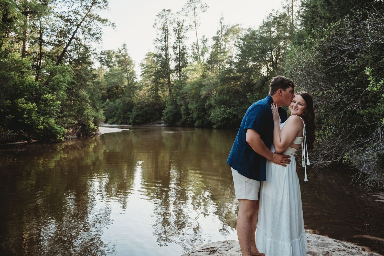 Romantic couple embracing and kissing near a calm woodland river surrounded by lush green trees.