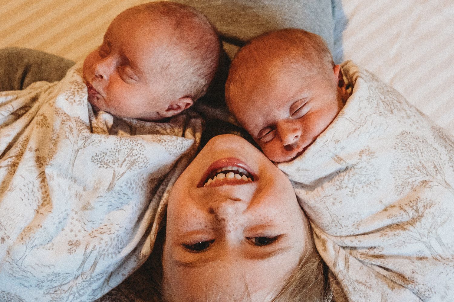 Overhead view of a smiling mother lying between two sleeping newborn twins wrapped in swaddles.