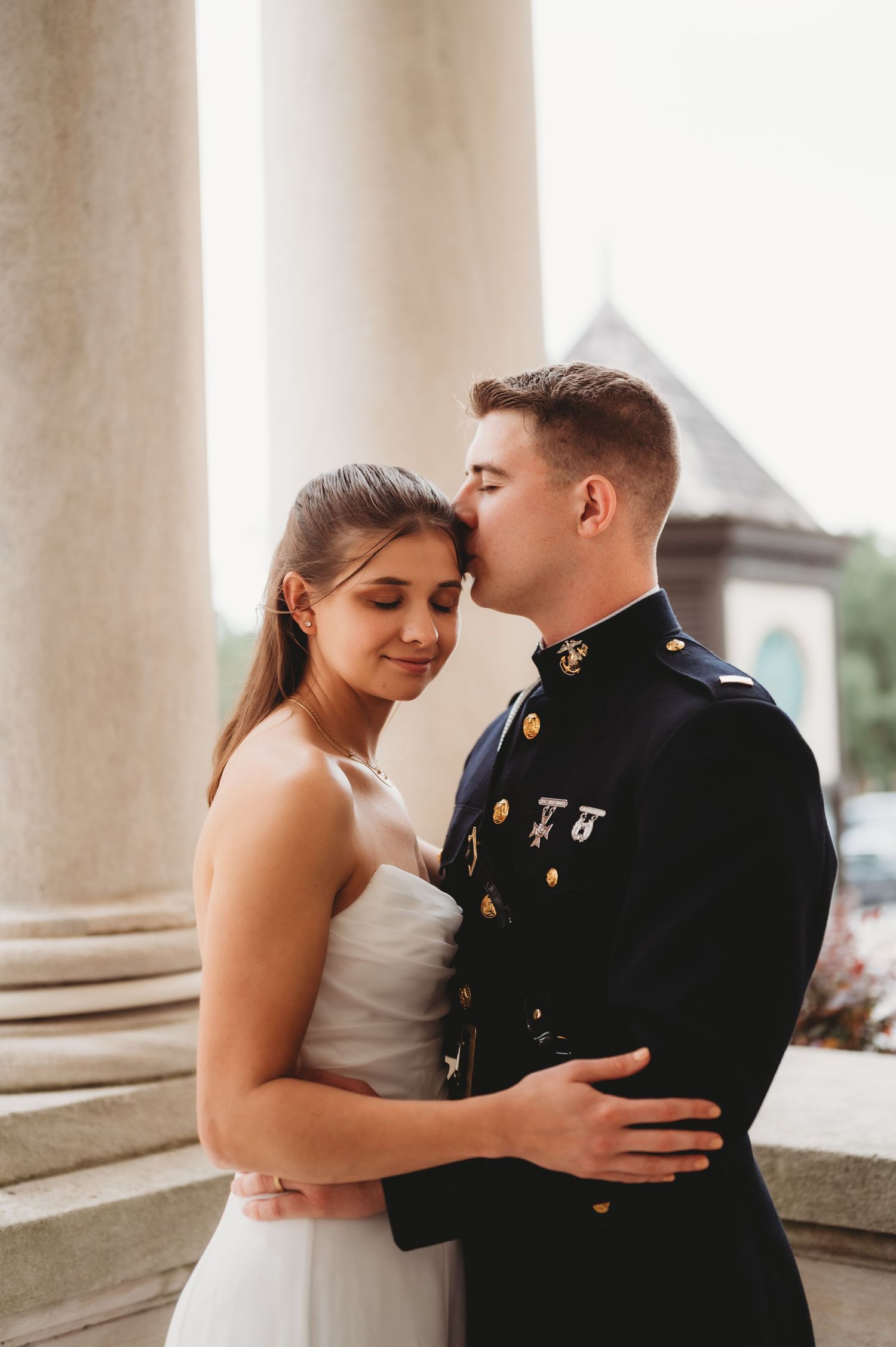 Marine groom kisses bride's forehead as she smiles softly near elegant stone columns.