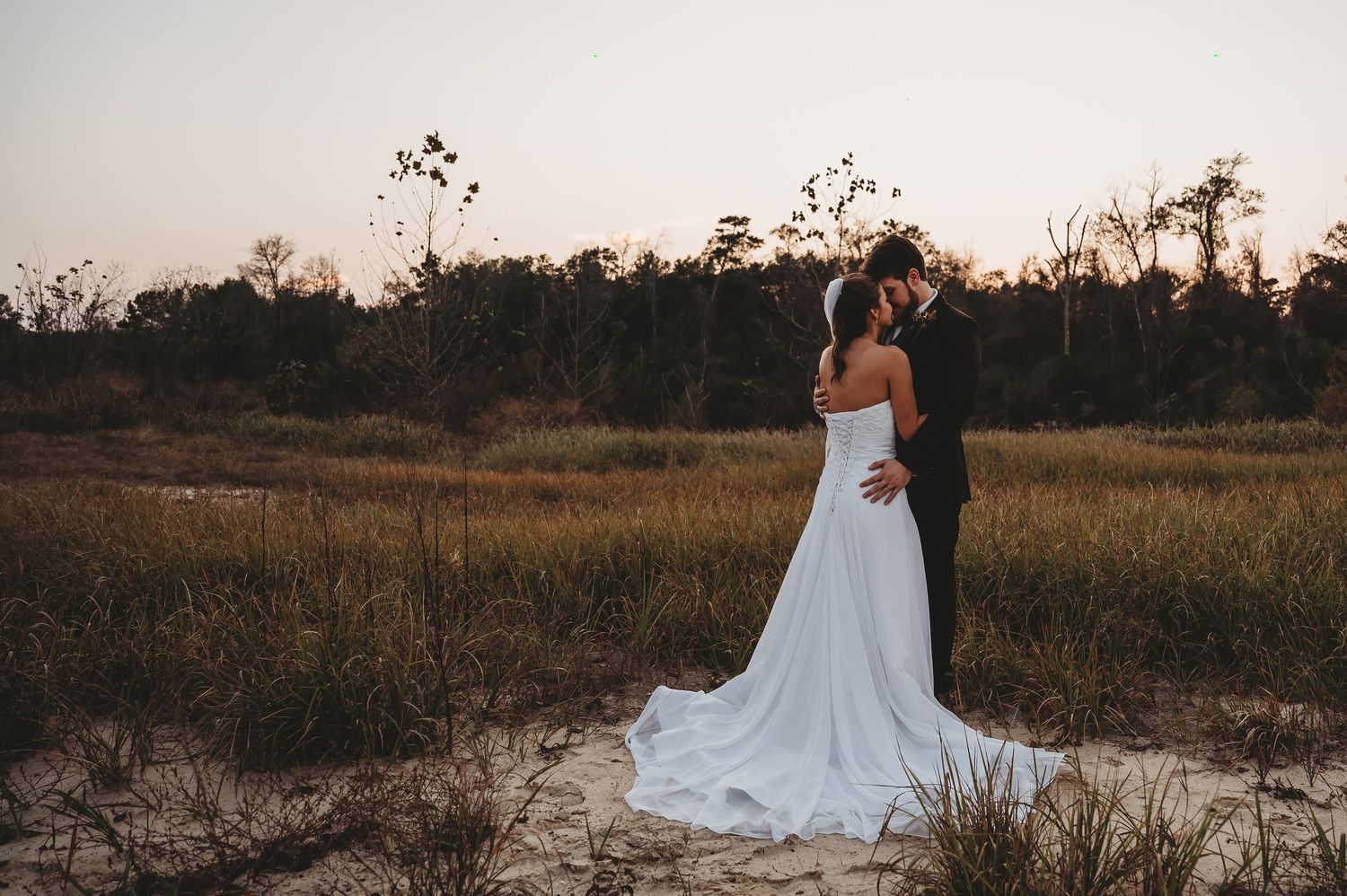 Bride and groom embrace at sunset in a rustic field, bride wearing a flowing white gown with train.