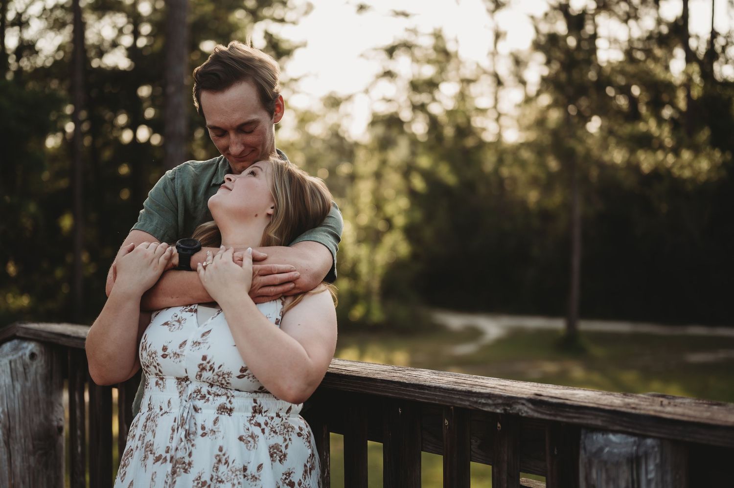 Couple embracing on wooden bridge surrounded by trees, man hugging woman in floral dress outdoors.