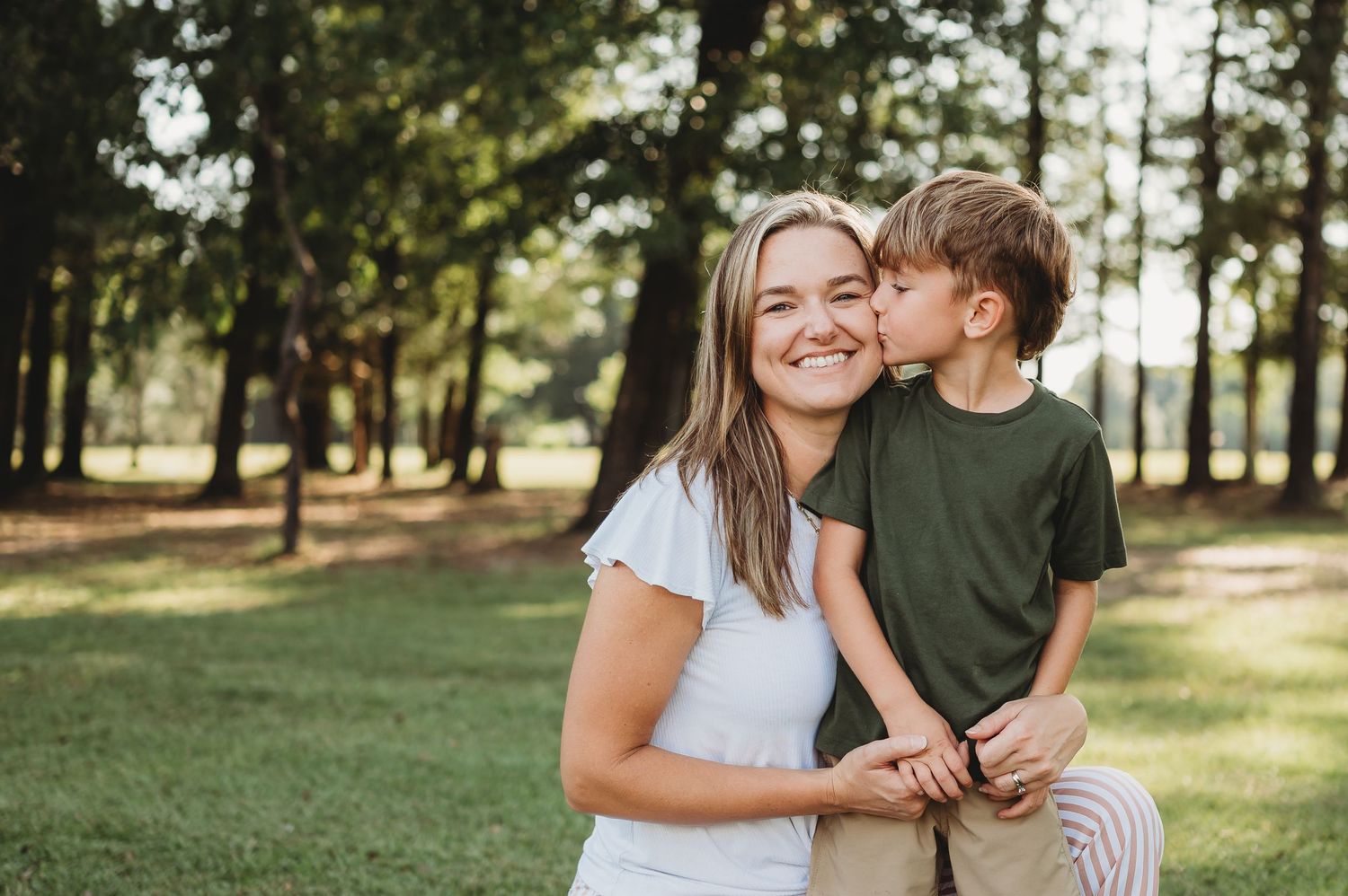 A smiling woman in a white shirt receives a kiss on the cheek from a young boy in a green shirt outdoors.