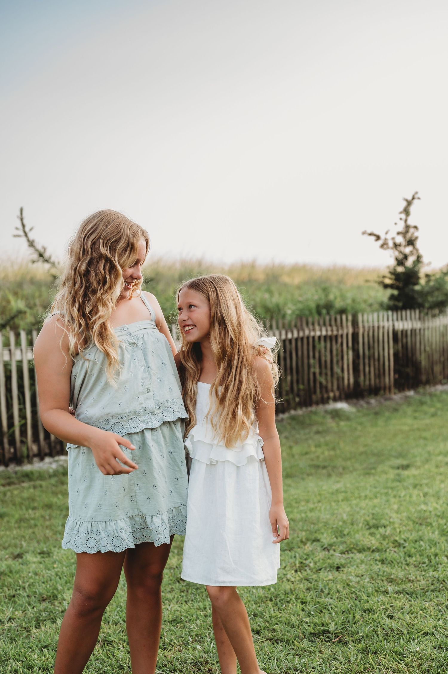 Two young girls with long blonde hair in summer dresses laughing together on a green lawn near a wooden fence.