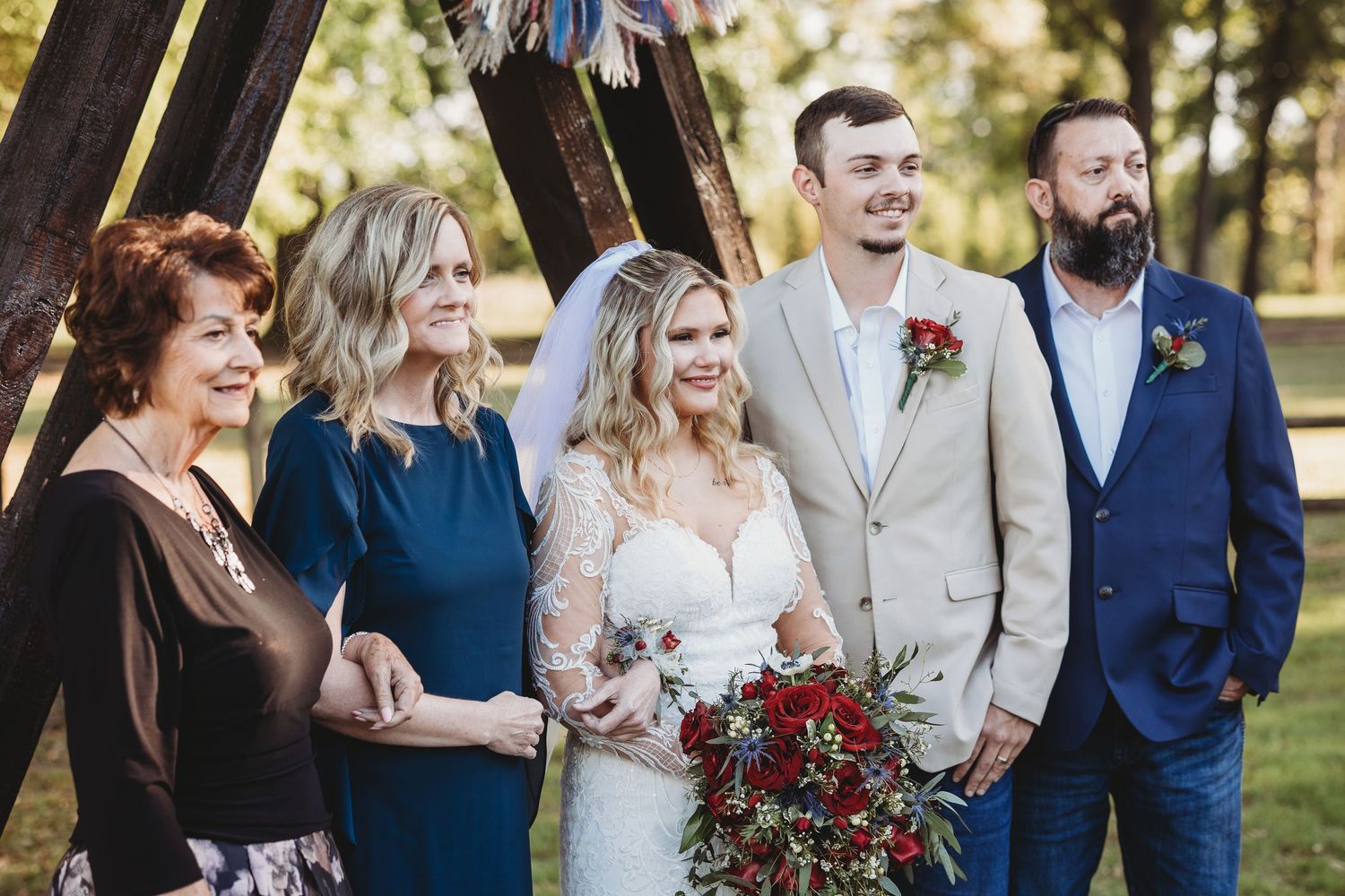 Wedding party group photo with bride holding red bouquet standing before a wooden teepee arch outdoors.