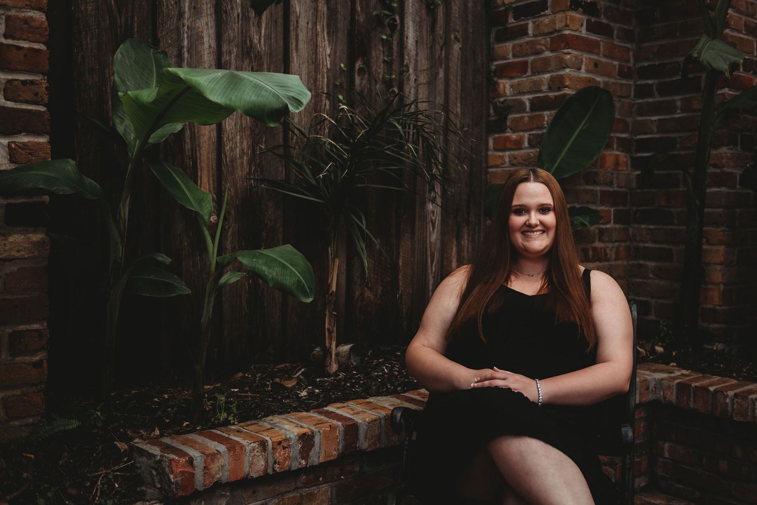 A young woman in a black dress sits on a brick ledge surrounded by tropical plants.