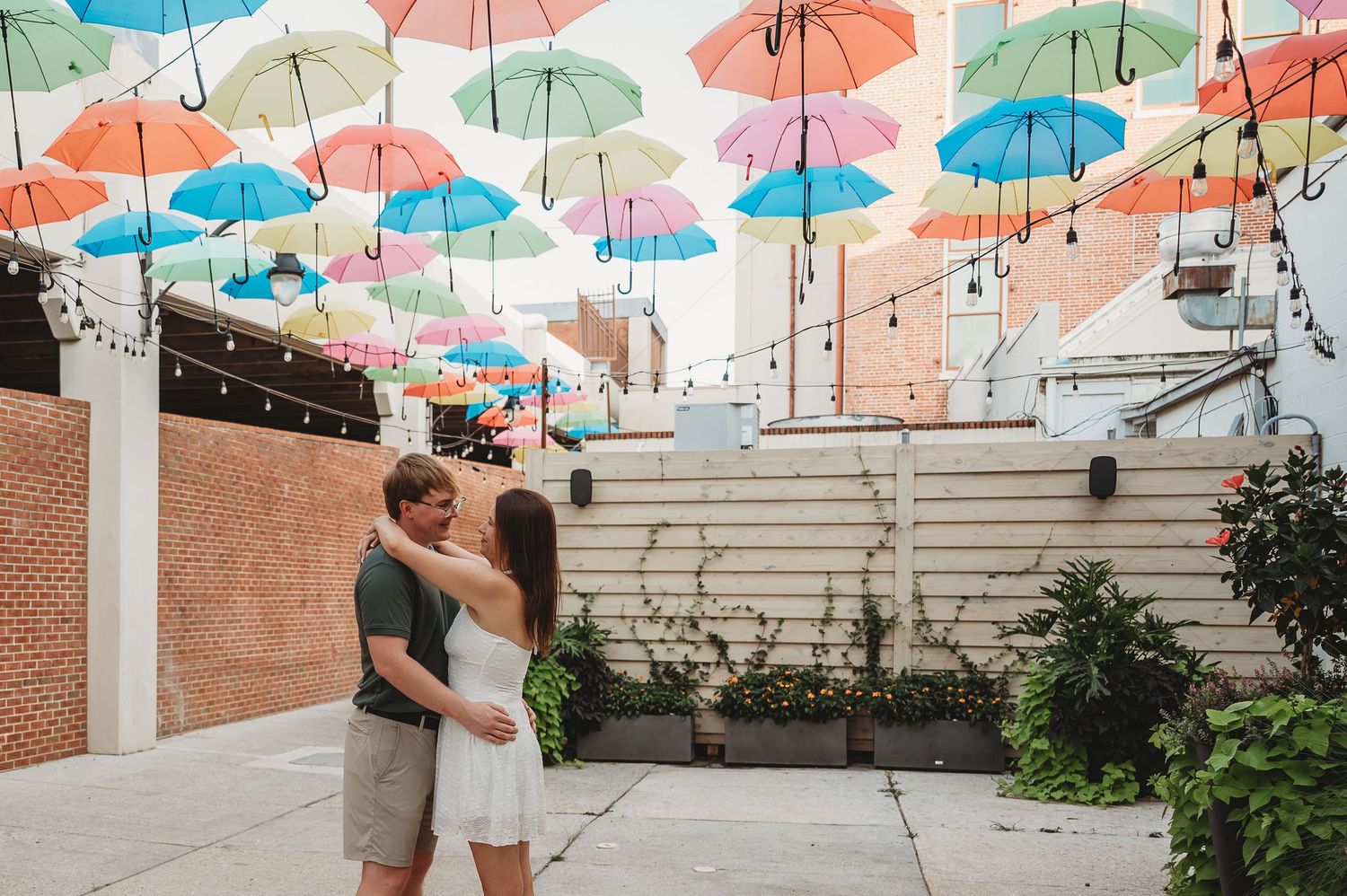 Couple sharing a romantic moment under colorful hanging umbrellas in a charming outdoor courtyard with string lights.