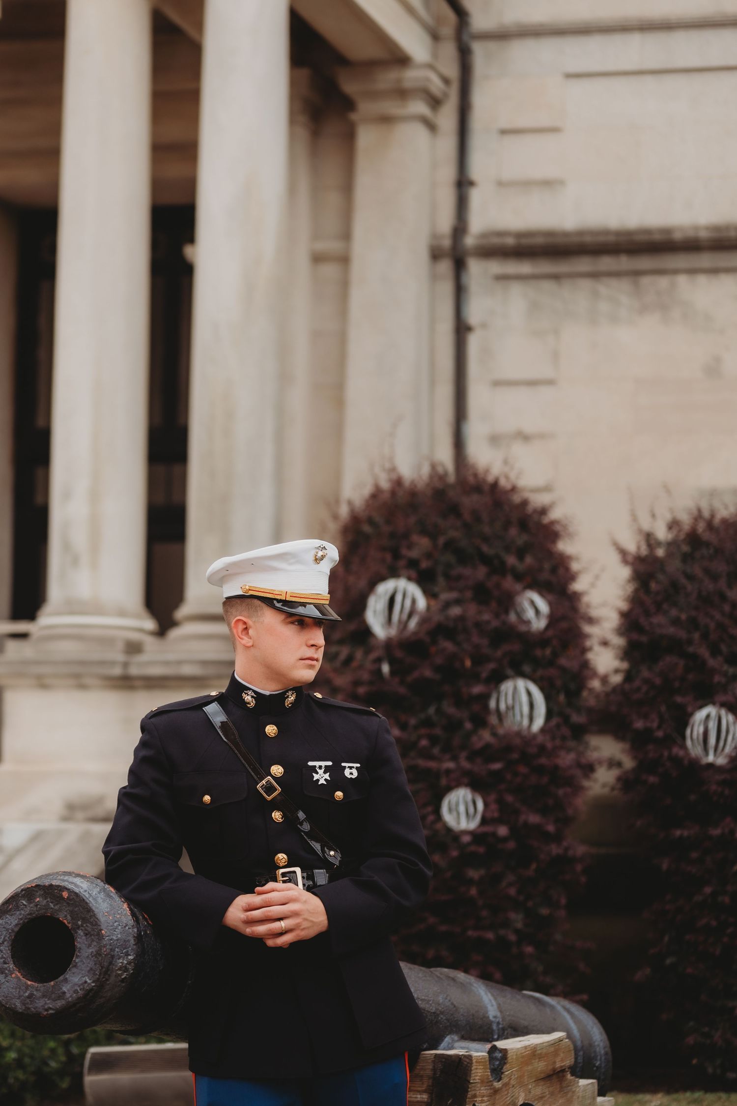A US Marine in dress blues uniform stands beside a historic cannon outside a neoclassical building.