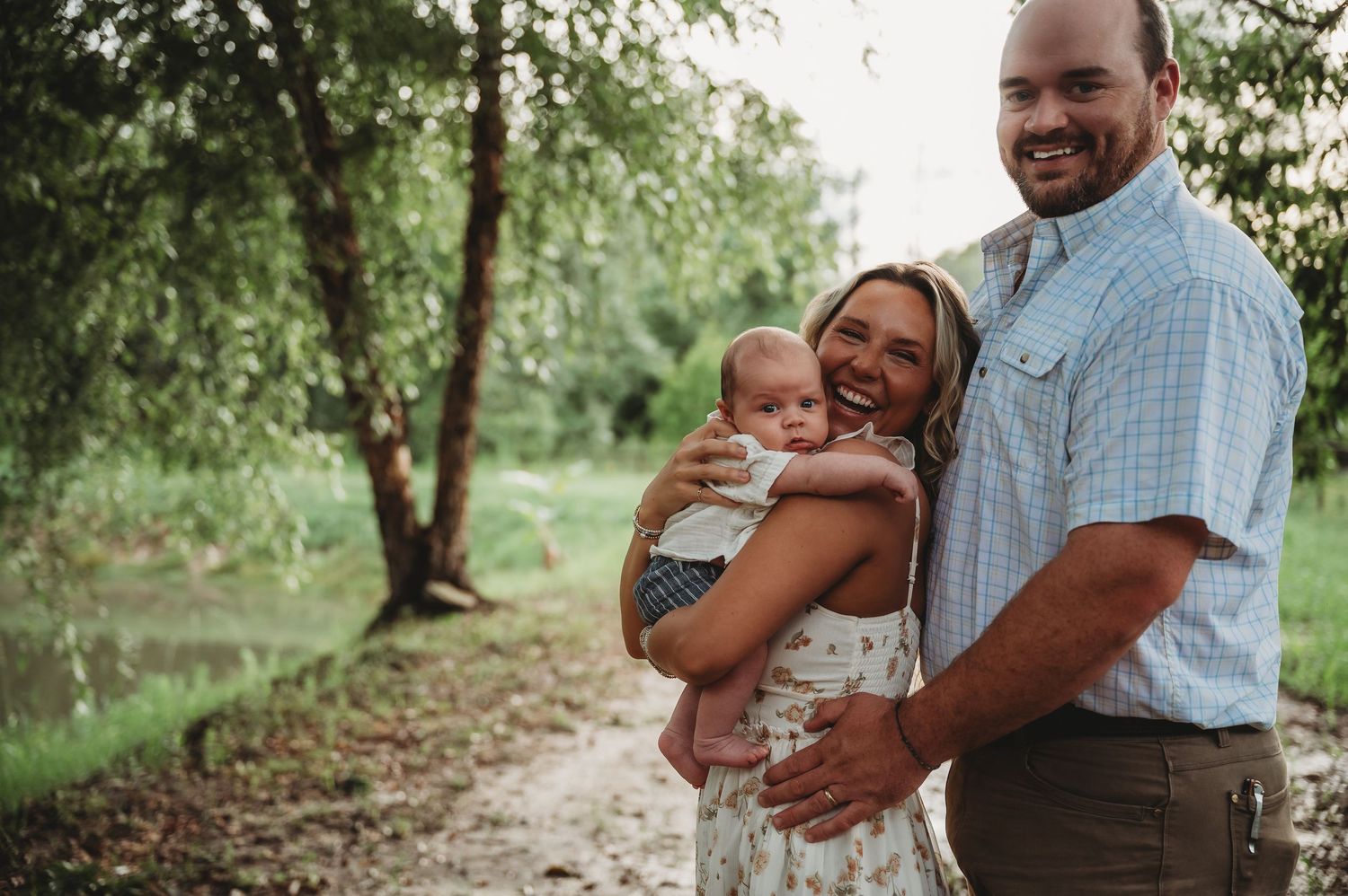 Happy family portrait outdoors, smiling couple holding baby near lush green trees on a sunny day.