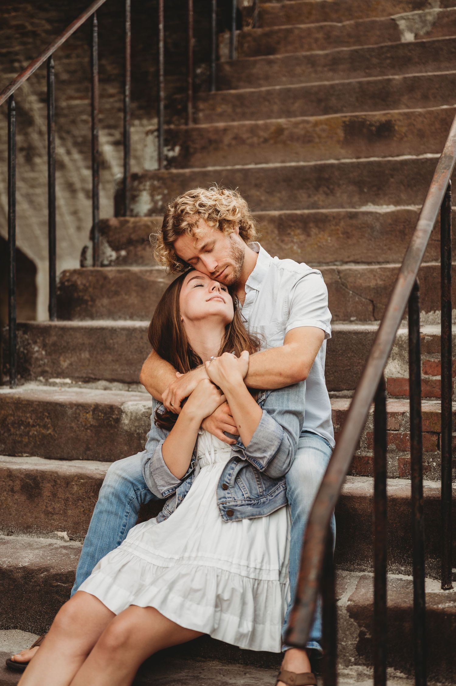 Couple sitting on brick steps embracing, man kissing woman's head, wearing casual denim and white dress.